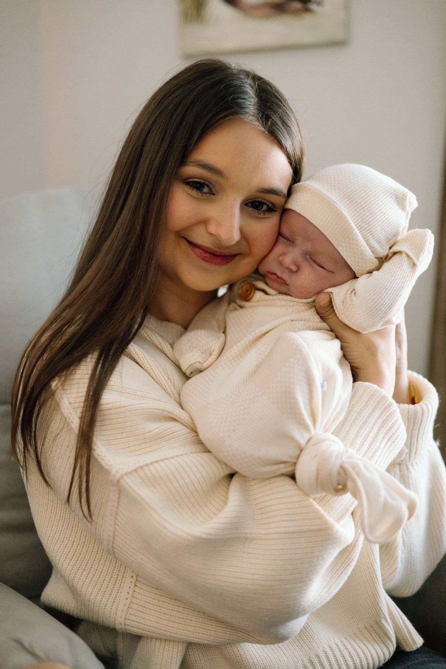 Brunette mother in cream sweater smiling while gently holding sleeping newborn baby against her cheek.