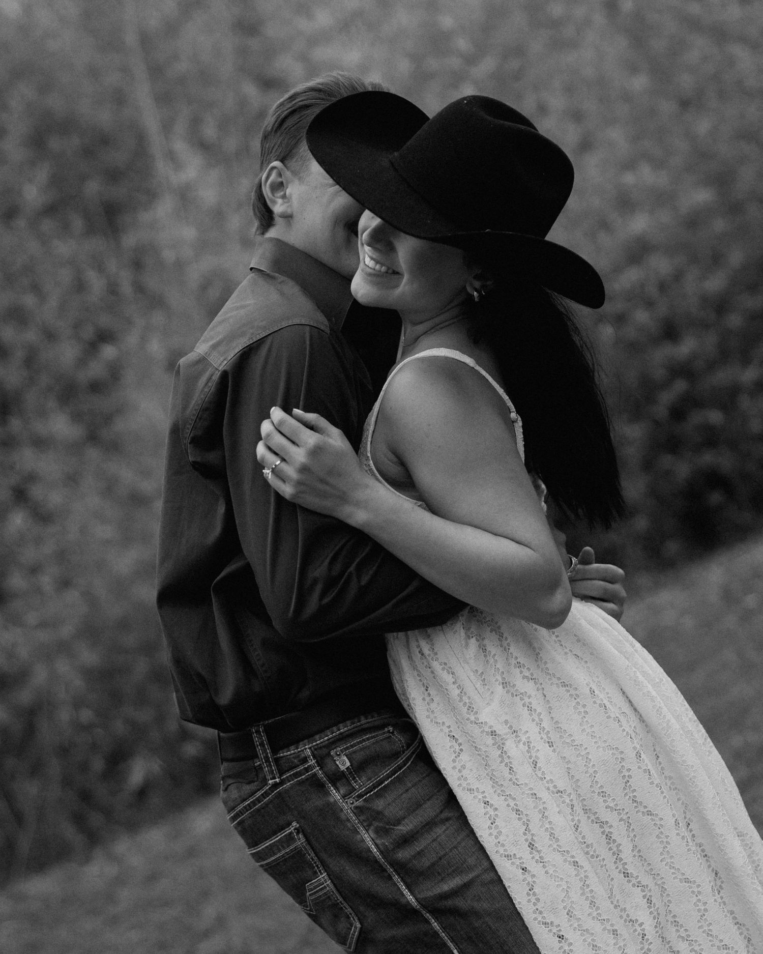 Black and white photo of couple embracing outdoors, woman wearing cowboy hat and lace dress.