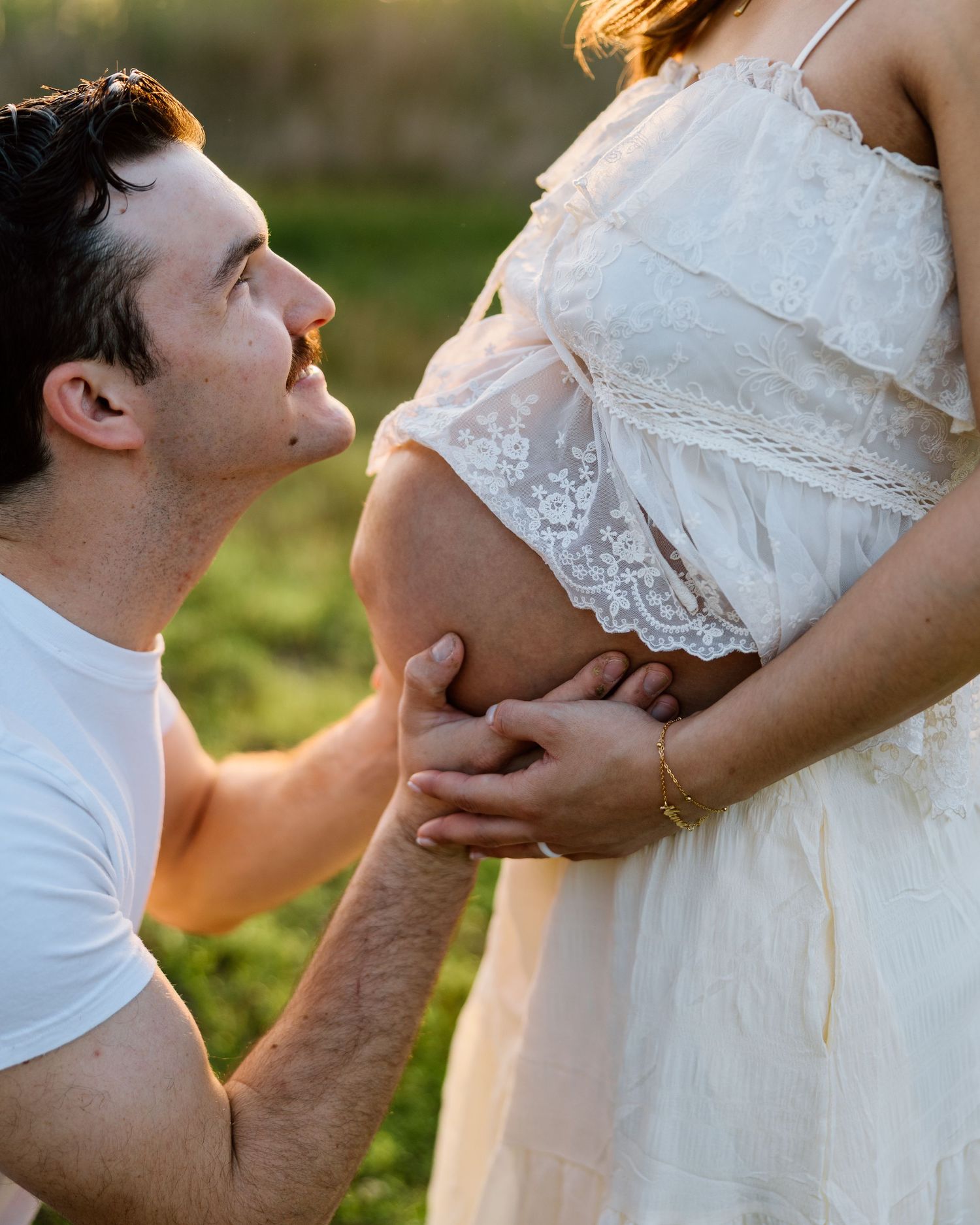 A man kneeling and holding a pregnant woman's belly outdoors in golden light, both dressed in white.