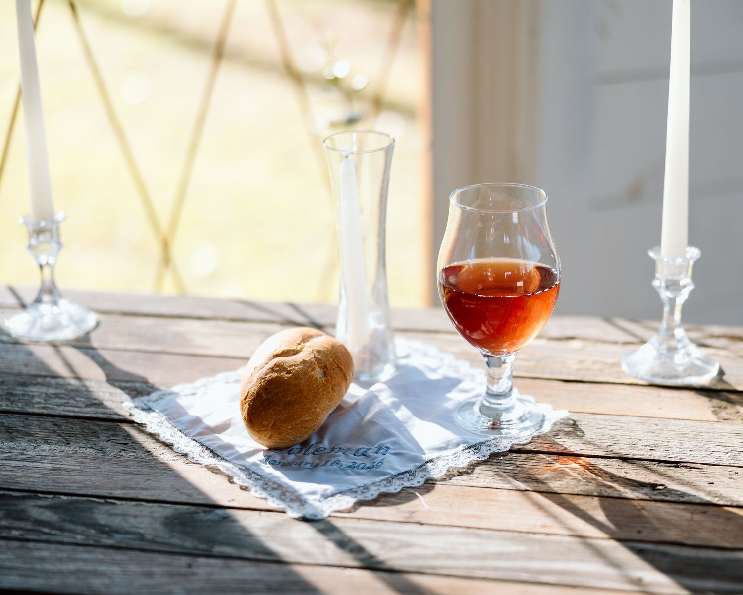 Rustic wooden table with a glass of amber wine, crystal decanter, bread roll, and candles in soft sunlight.