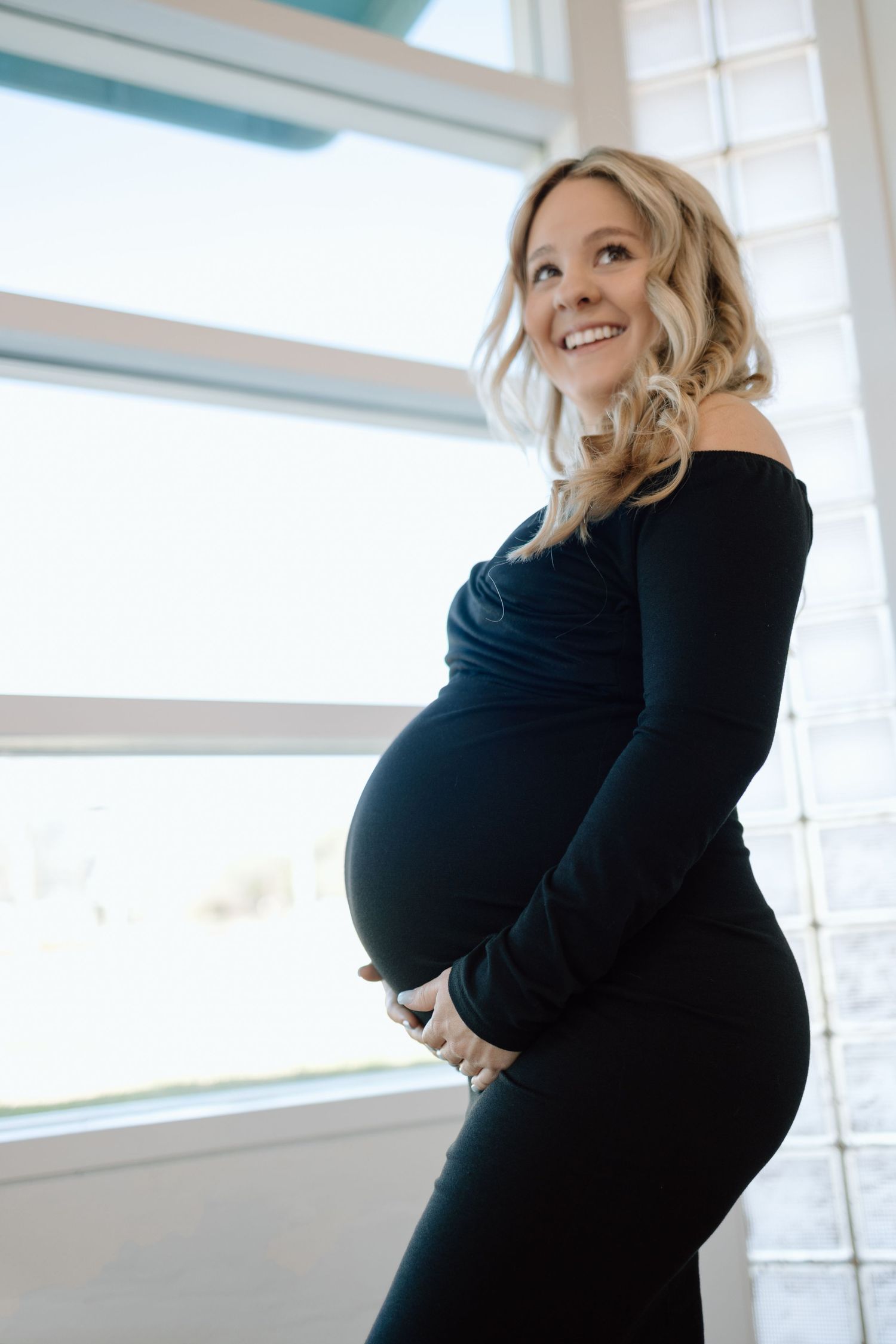 A pregnant woman in a fitted black dress stands by bright windows, smiling while cradling her baby bump.