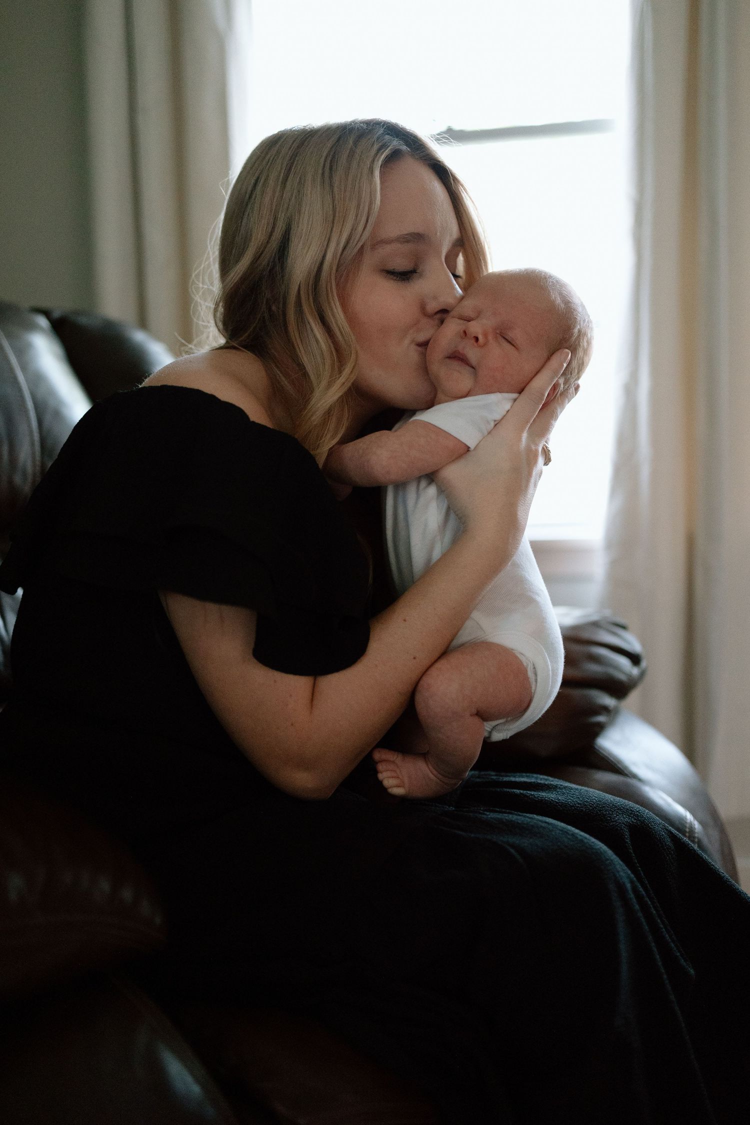 A tender moment of affection as someone in black holds and kisses a swaddled newborn baby on a leather couch.