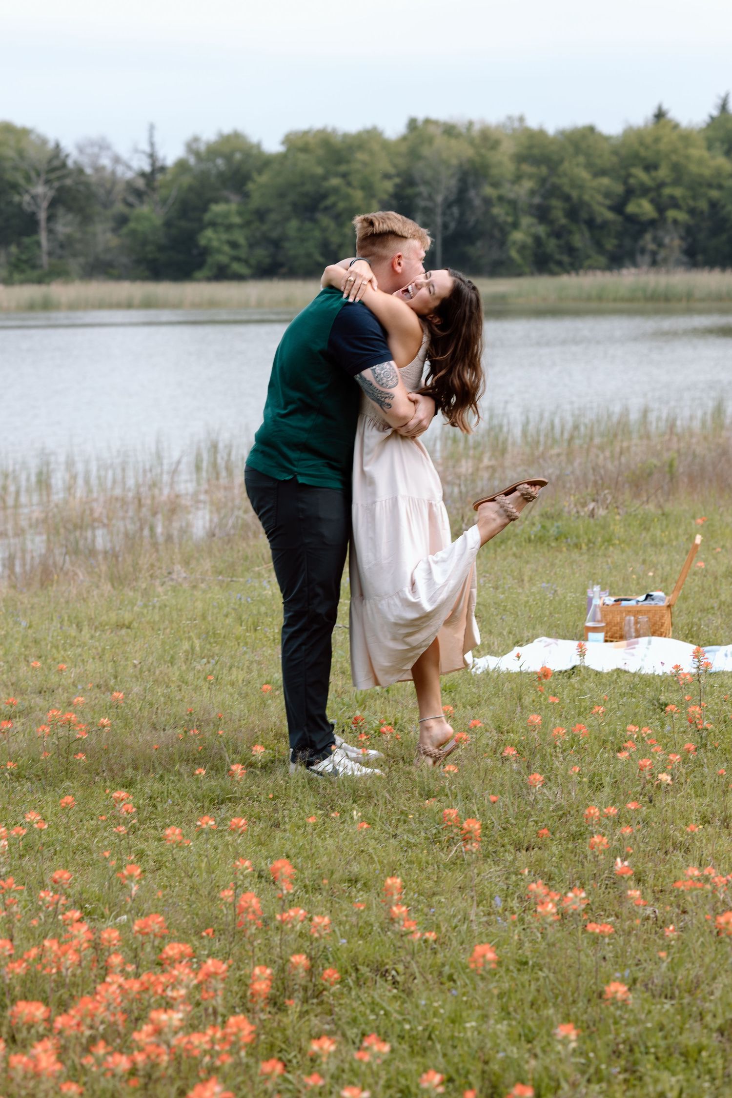 A romantic couple embraces in a field of orange wildflowers beside a lake with a picnic setup.