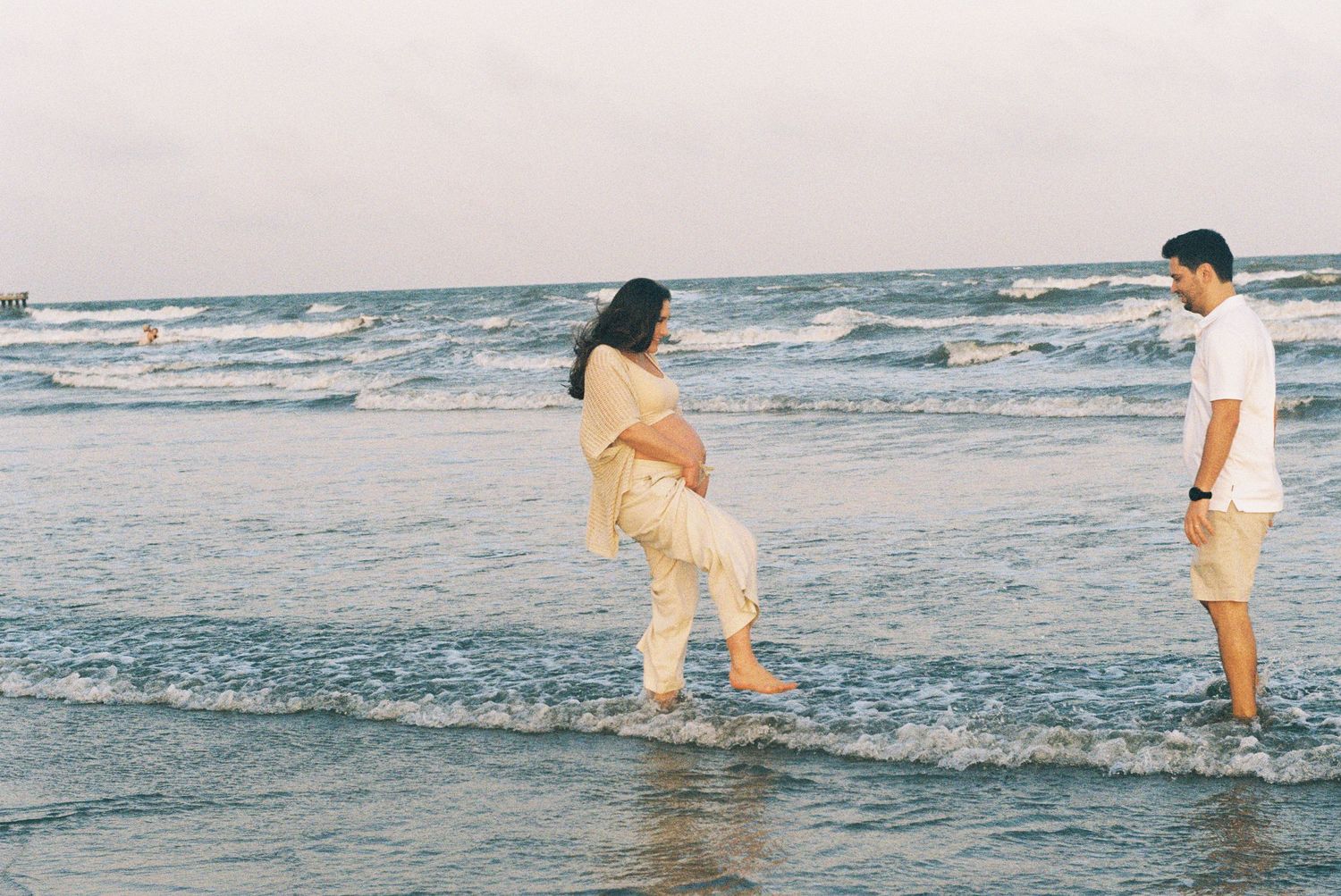 A couple plays in the ocean waves during a beach maternity photoshoot.