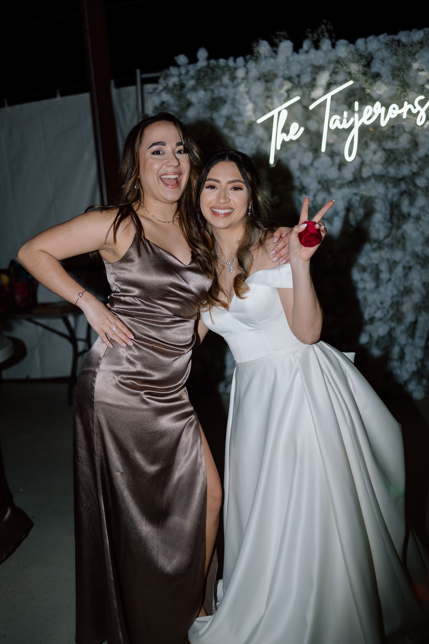 Wedding celebration with bride and friend posing in front of illuminated neon sign and floral wall.