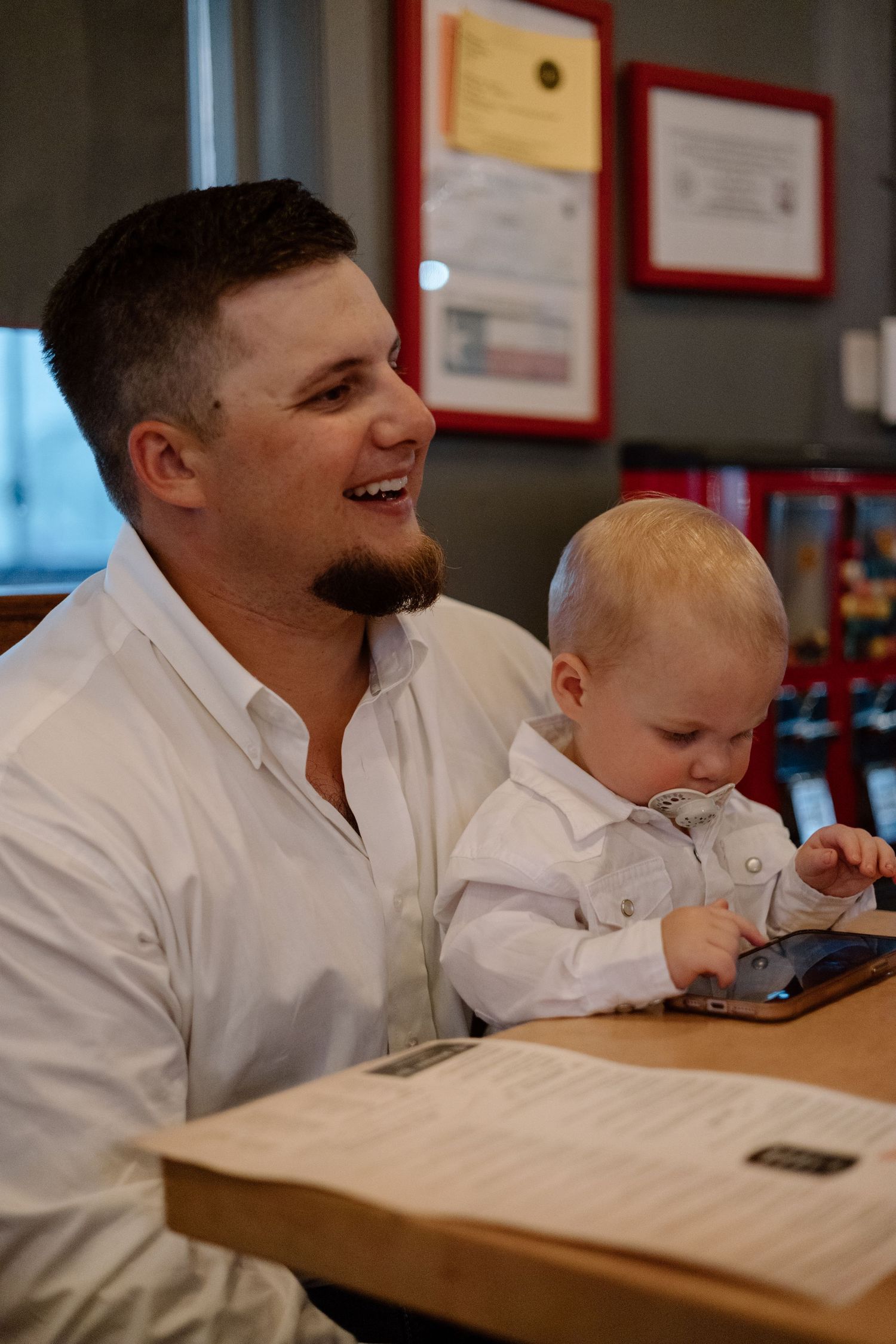 Smiling man in white shirt holds baby playing with tablet at restaurant table with framed certificates on wall.