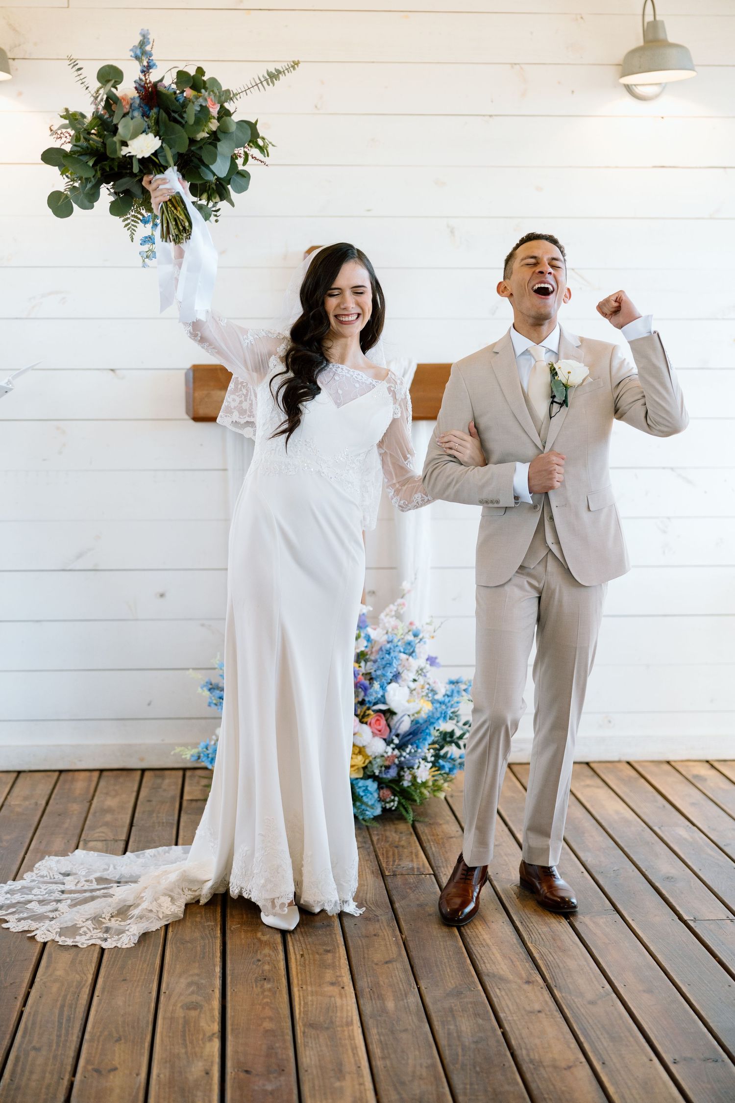 Joyful bride raises bouquet and groom pumps fist celebrating after wedding ceremony in white barn venue.