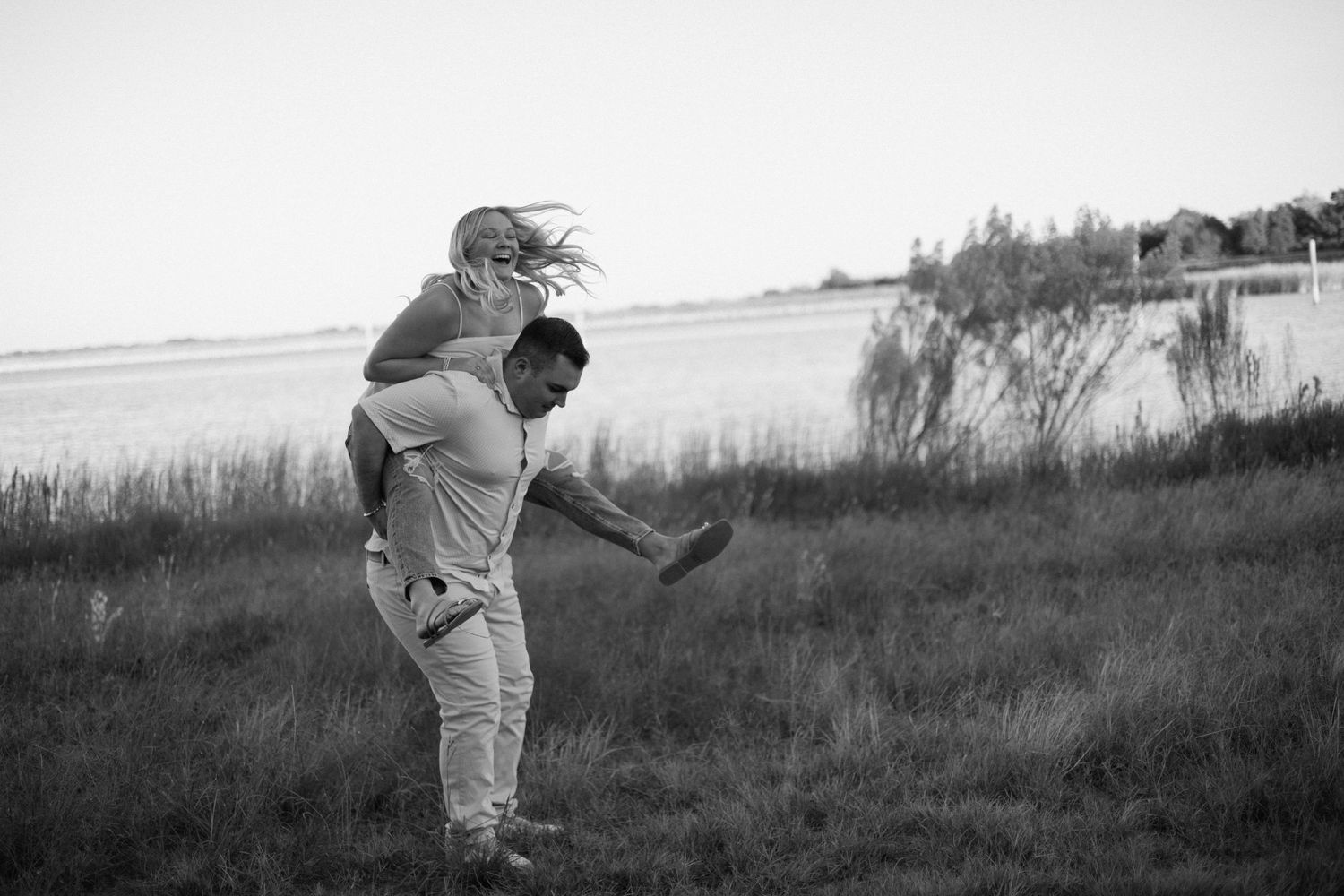 Black and white photo of a man giving a woman a piggyback ride in a grassy field near a lake.