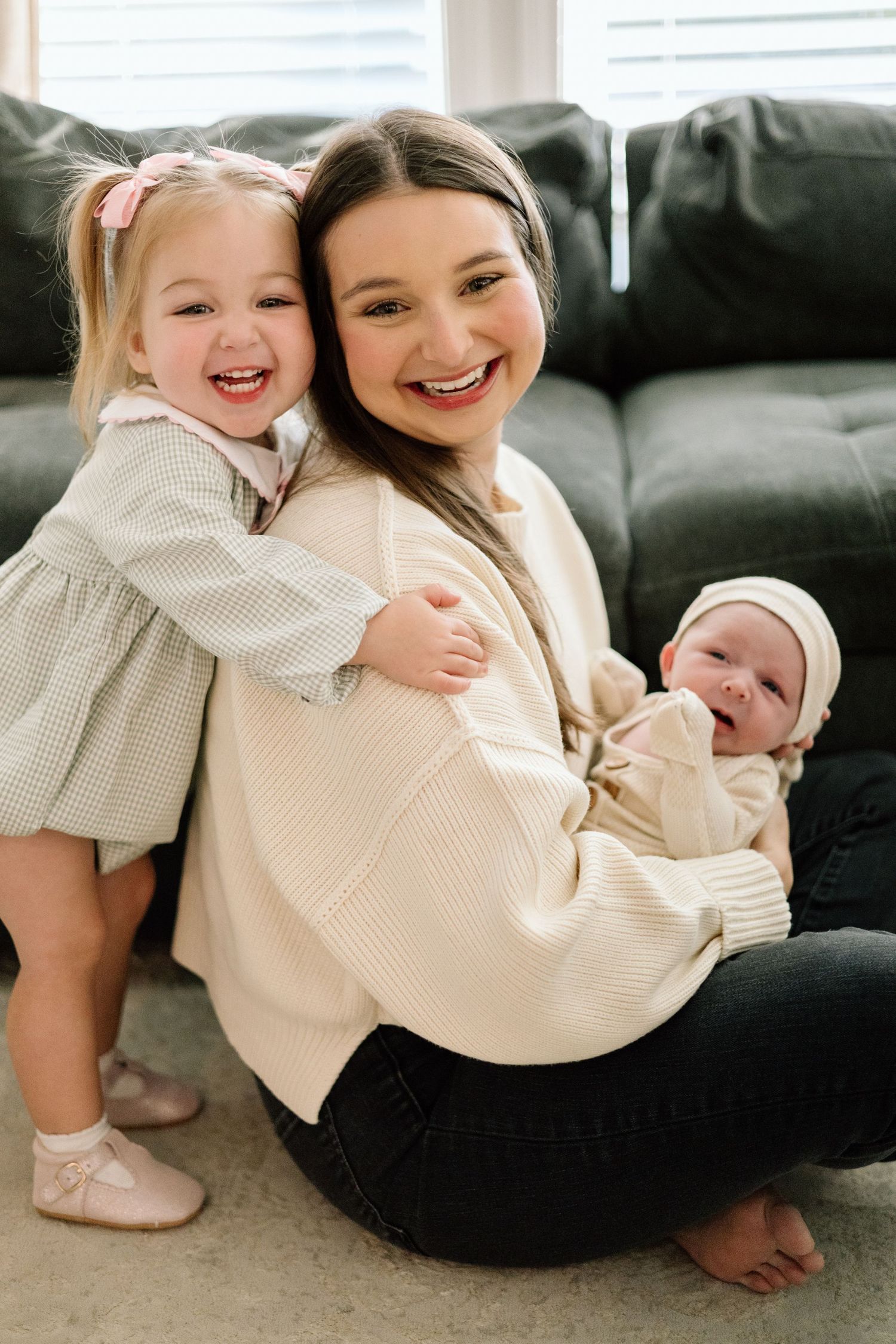 Smiling mom sitting on floor holding newborn baby while toddler girl in pink bow hugs her from behind.