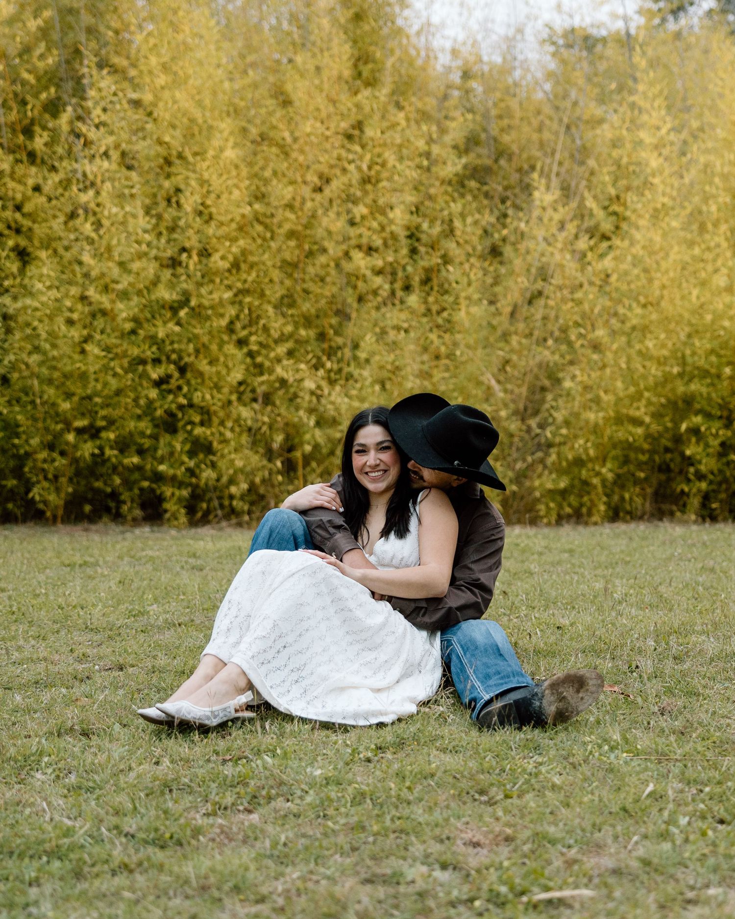 Couple sitting on grass, man in cowboy hat kissing woman in white dress near golden trees.
