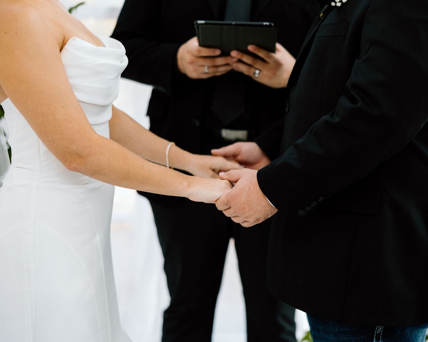 Bride and groom holding hands during wedding ceremony while officiant reads from tablet in background.