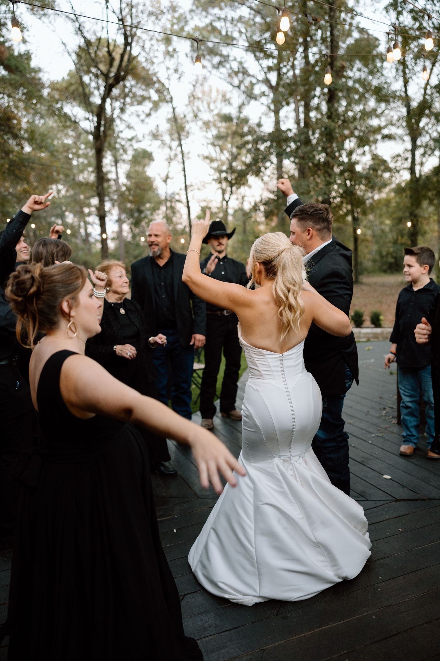 Bride in white mermaid gown dances with groom outdoors at wedding reception surrounded by guests in black attire.