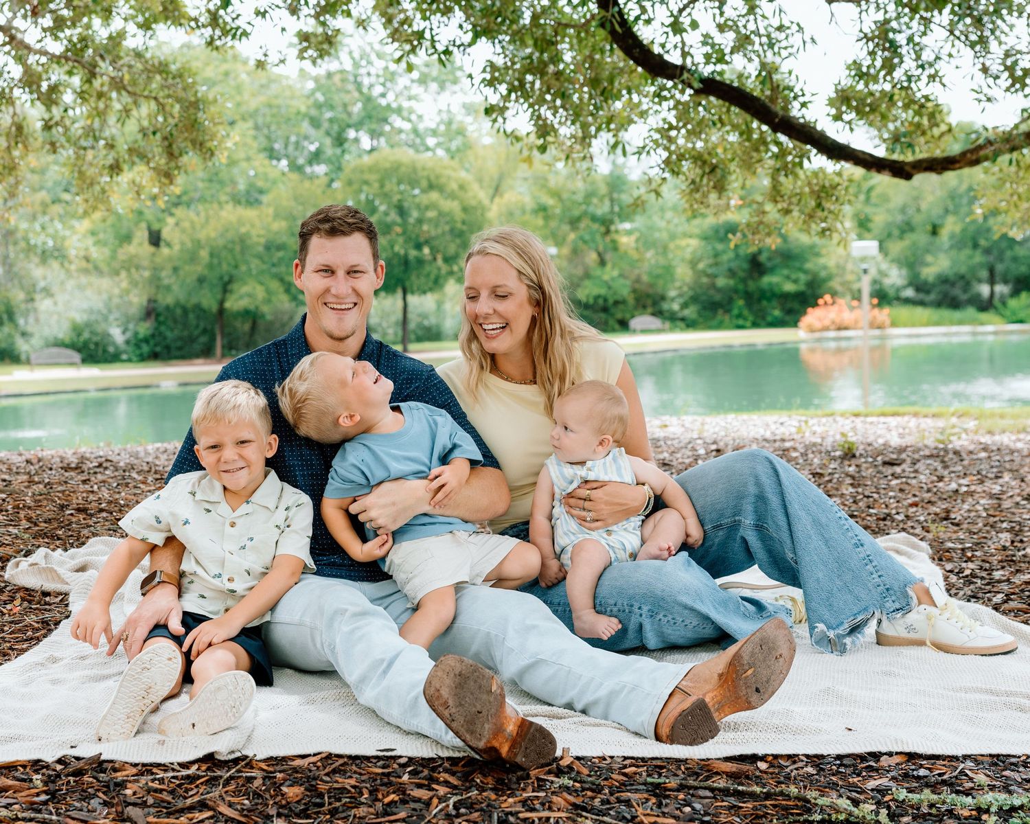 Happy family of five sitting on a blanket outdoors near a pond, parents smiling with three young children.