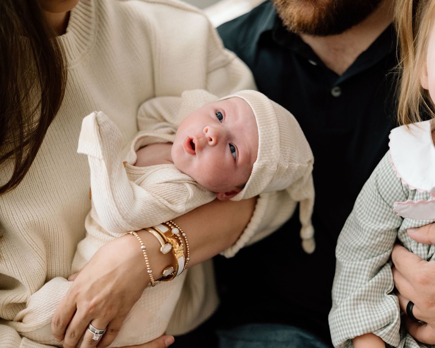 Newborn baby in white knit hat cradled in parent's arms, wearing cream outfit, with family members nearby.