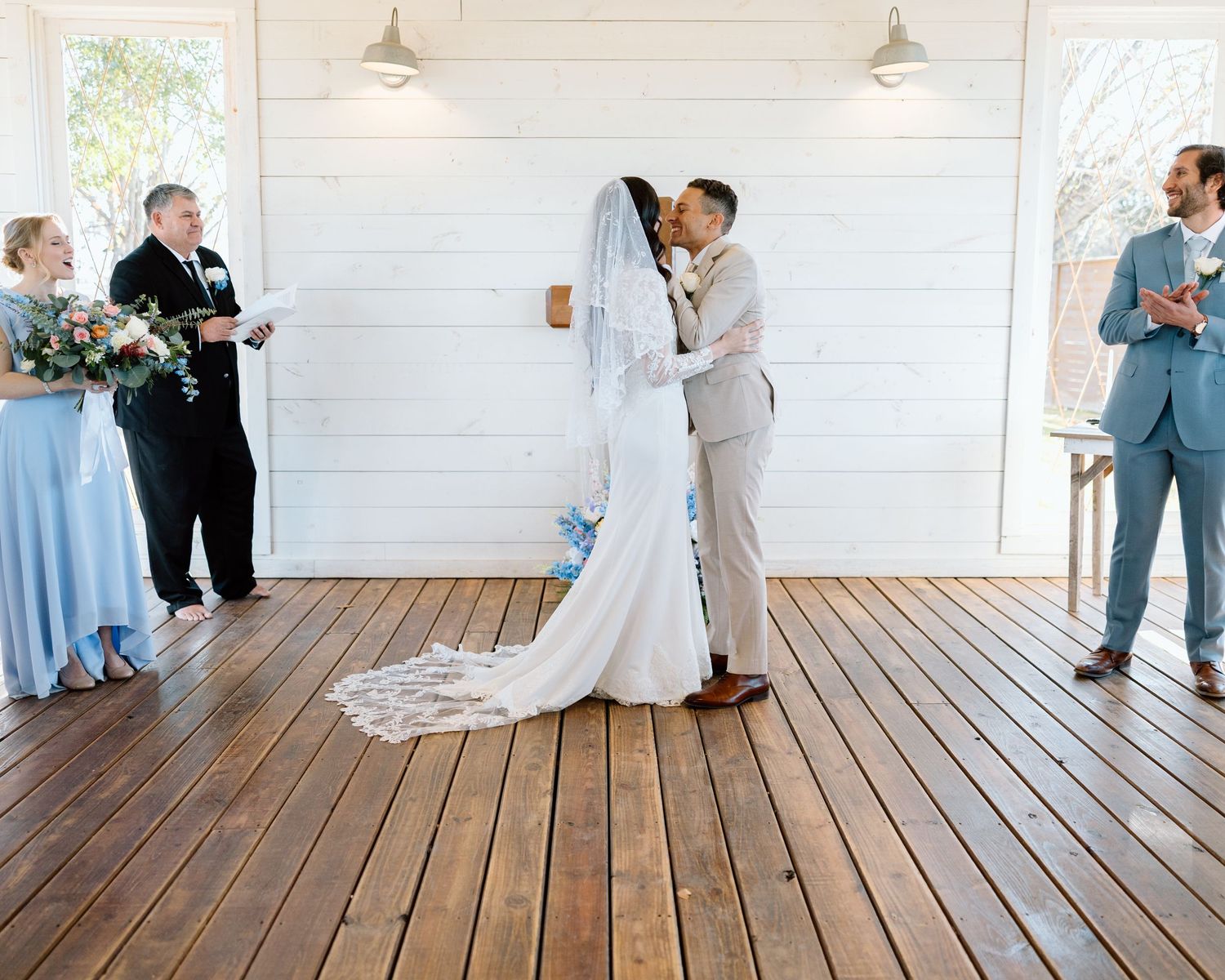 Newlyweds share first kiss at rustic white barn wedding venue with guests watching.