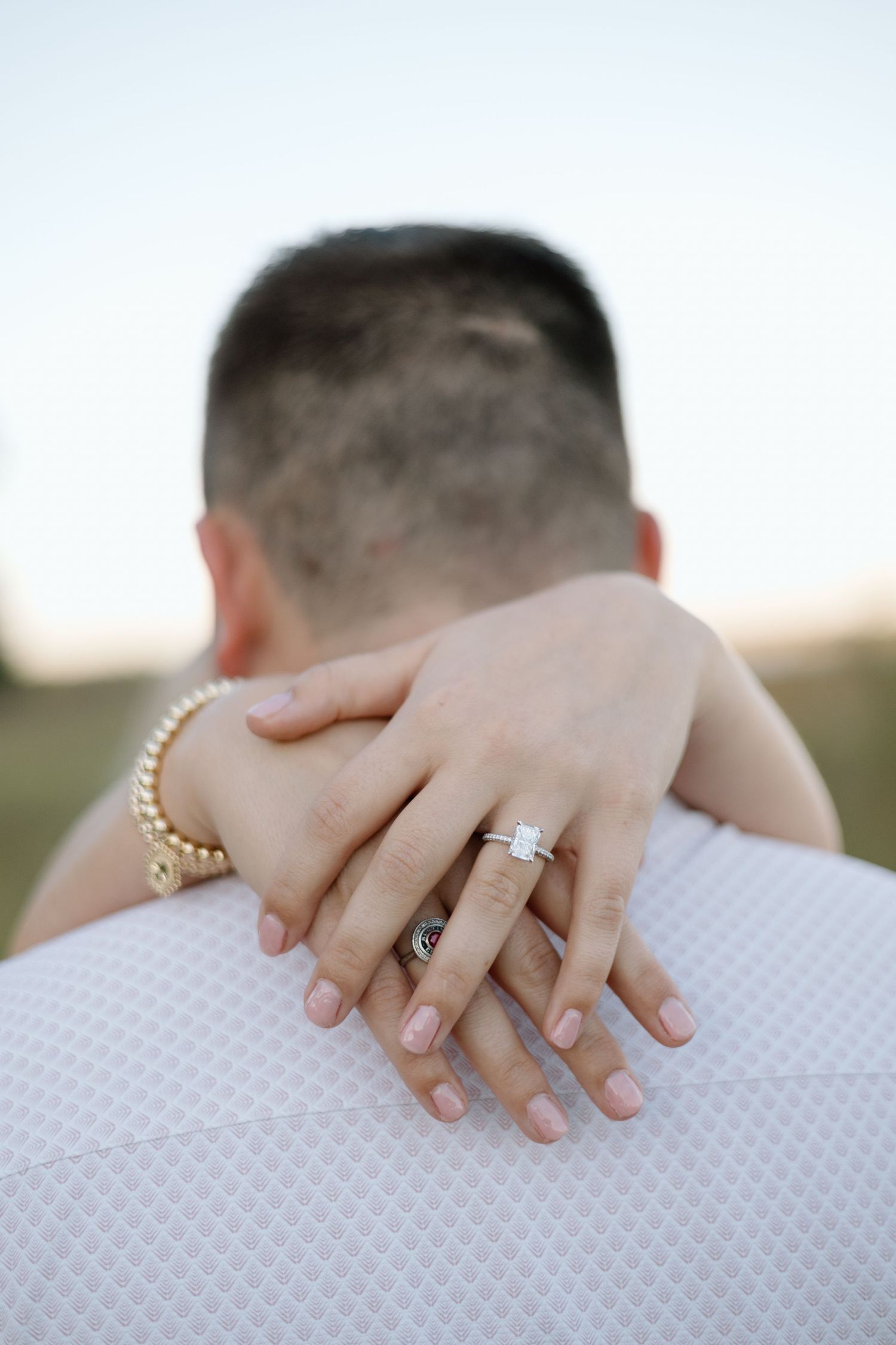 Close-up of a woman's hand with an engagement ring and gold bracelet resting on a man's shoulder.