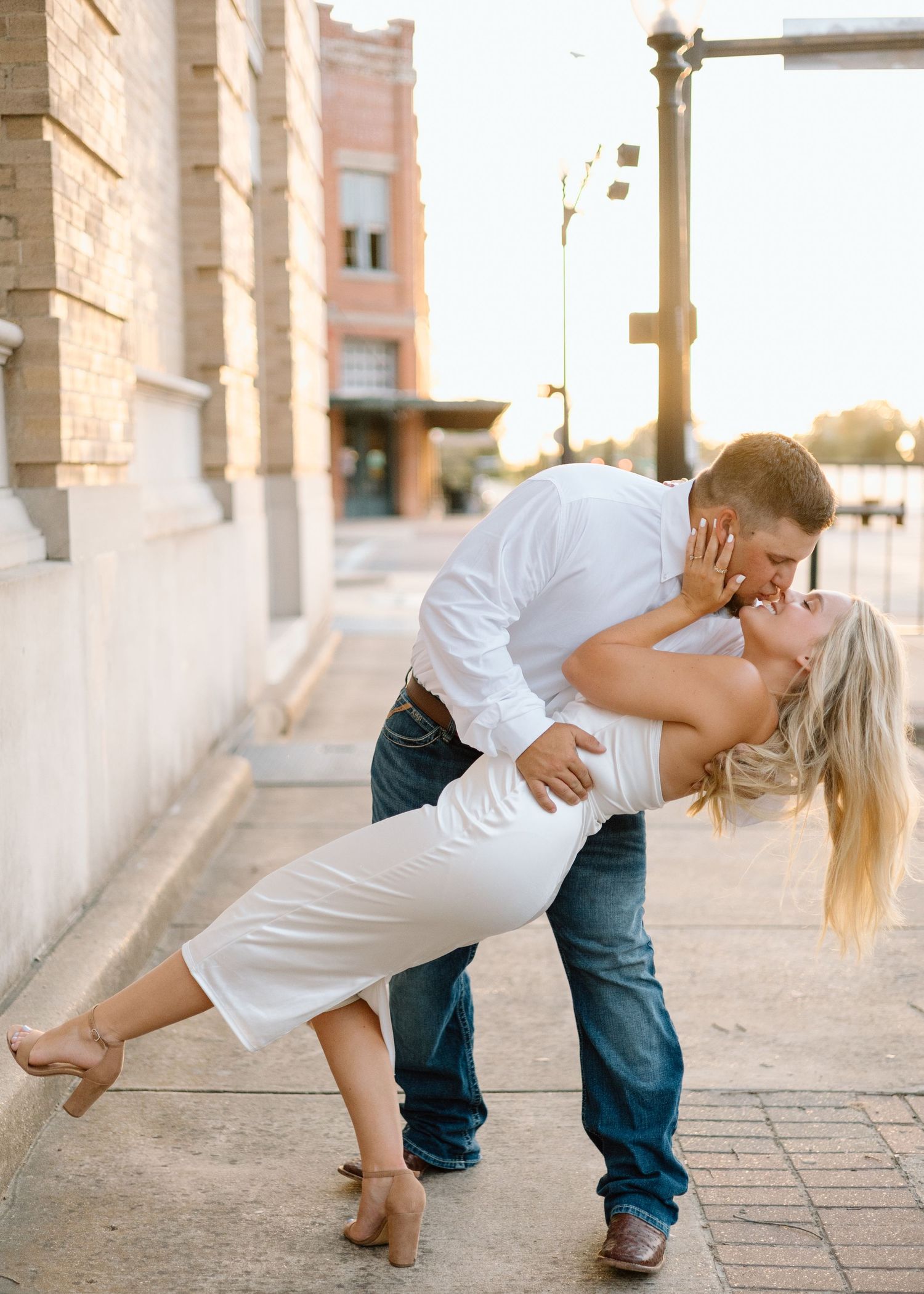 Couple sharing a romantic dip kiss on a sunny urban sidewalk, man in white shirt and jeans, woman in white dress.