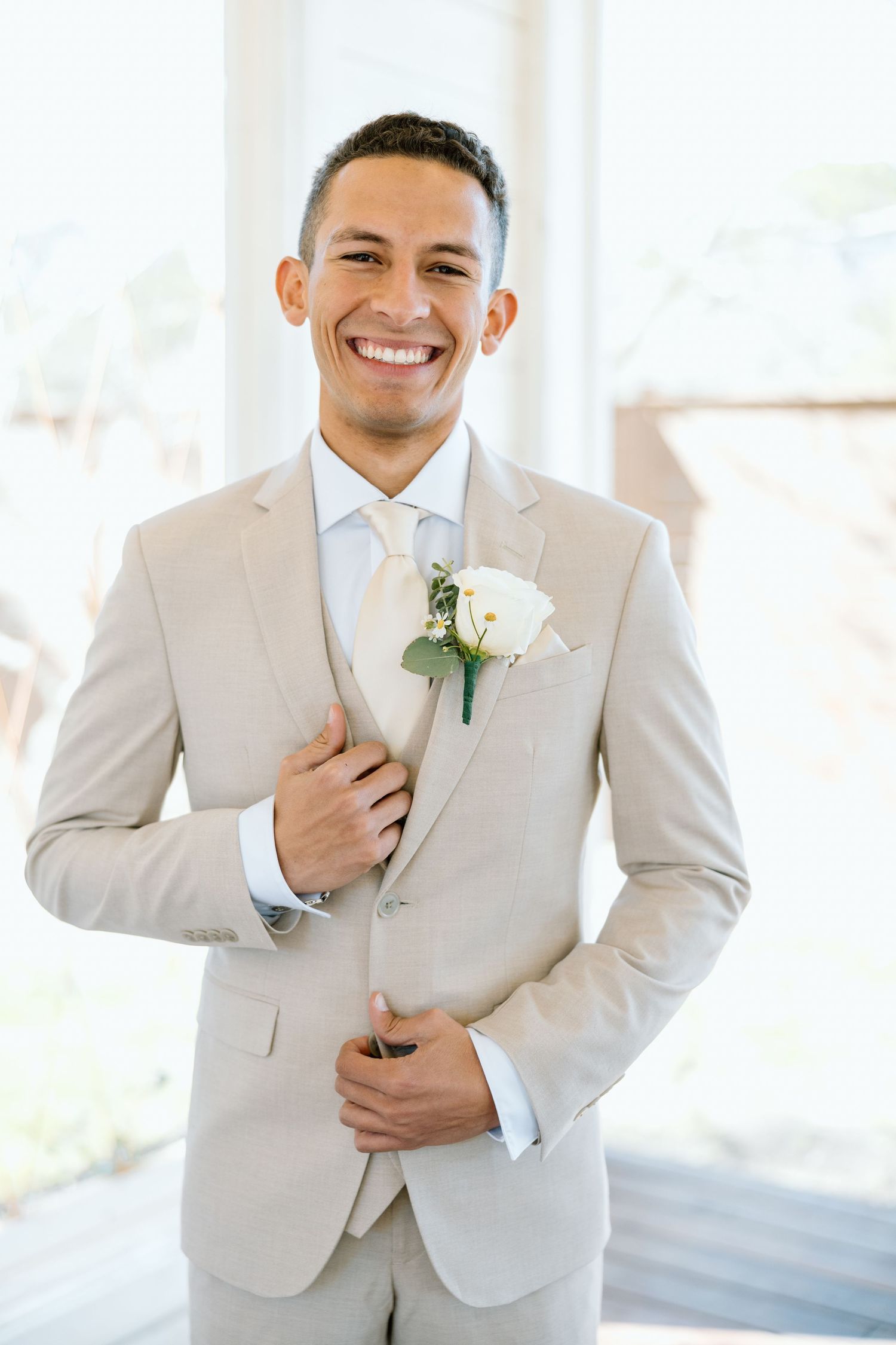 Smiling groom in beige suit adjusting lapel with white rose boutonniere indoors.