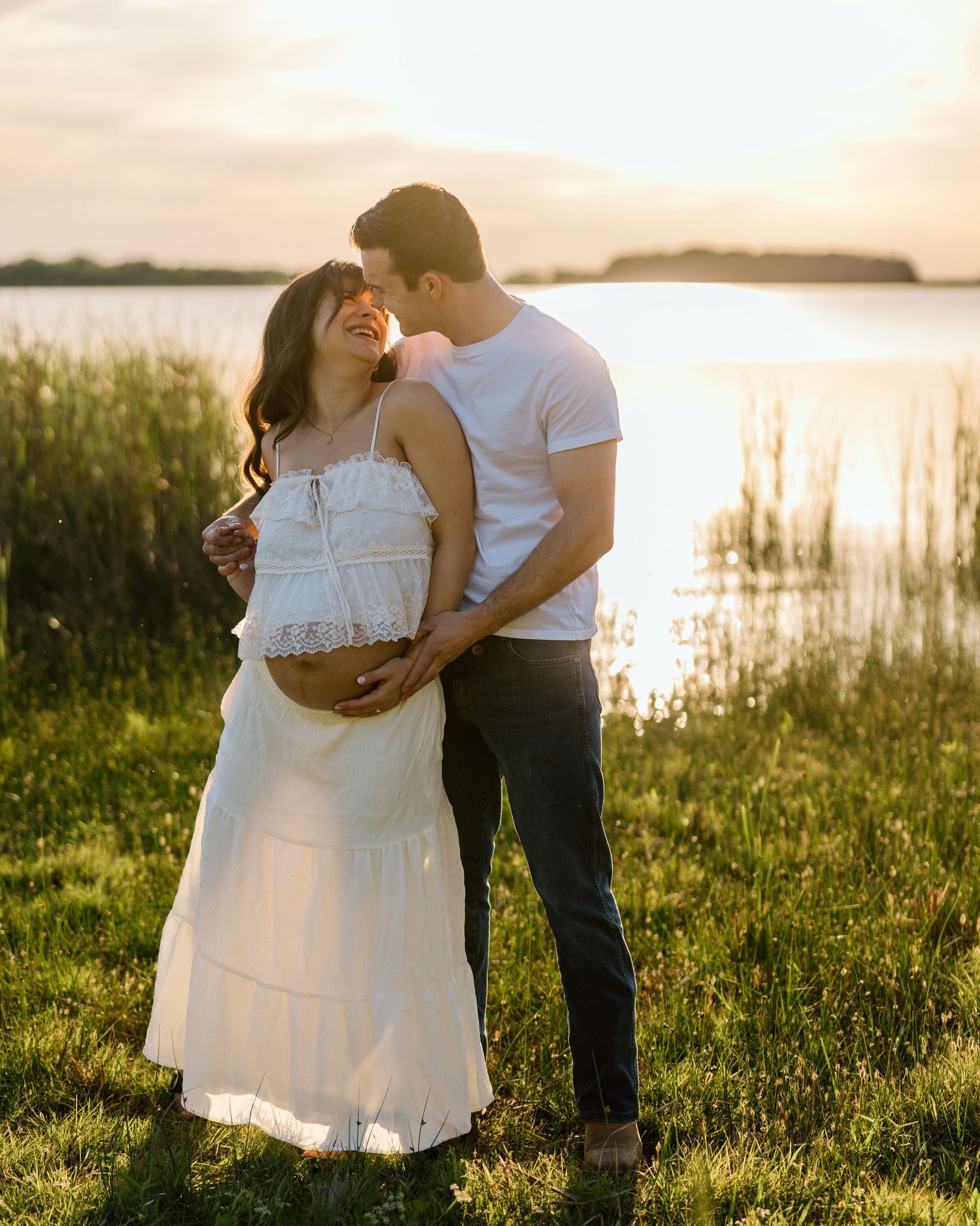 Couple embracing at golden hour by a lake, pregnant woman in white crop top and skirt, man in white tee and dark jeans.