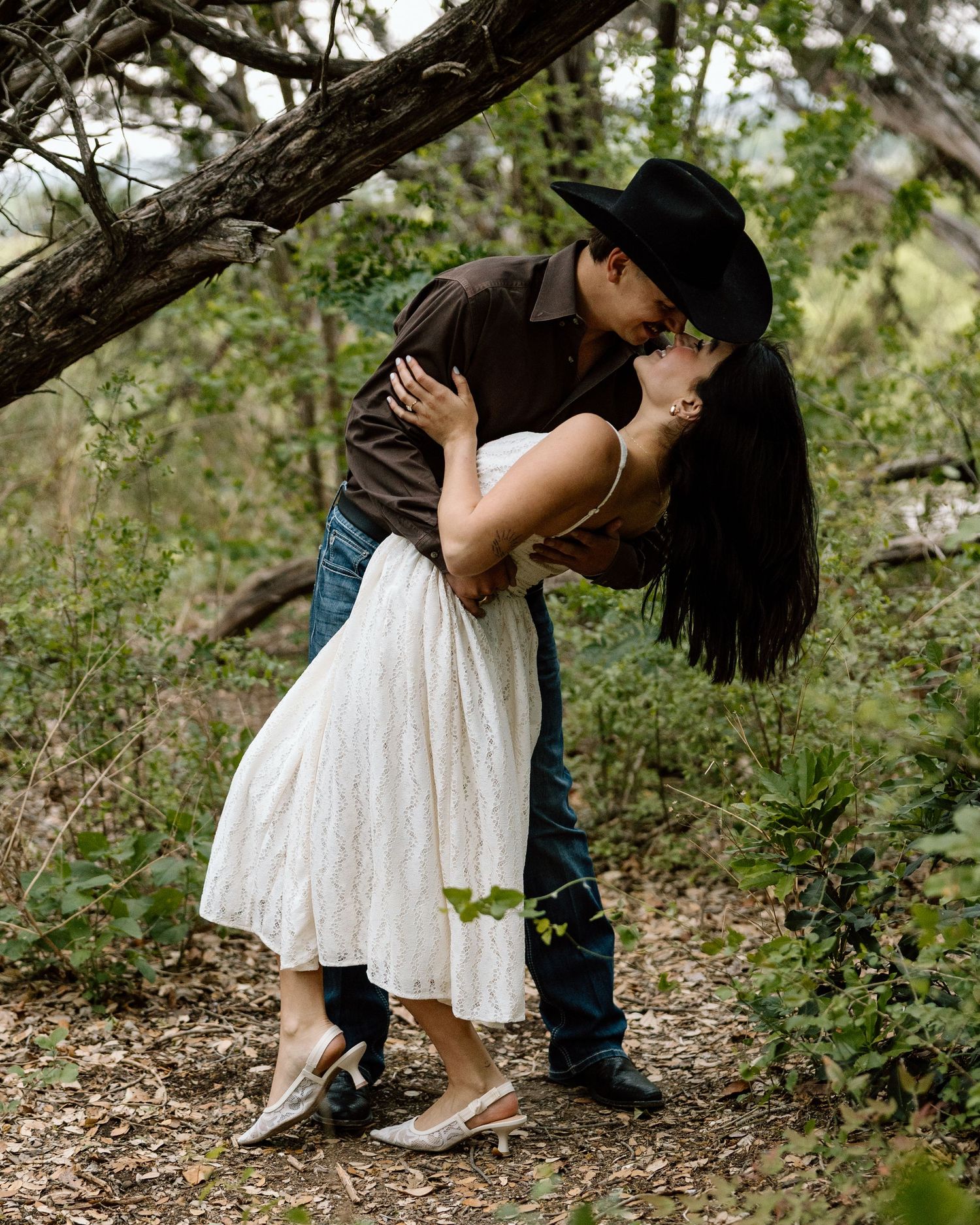 Couple sharing a romantic dip kiss outdoors, man in cowboy hat and woman in white dress, surrounded by lush greenery.
