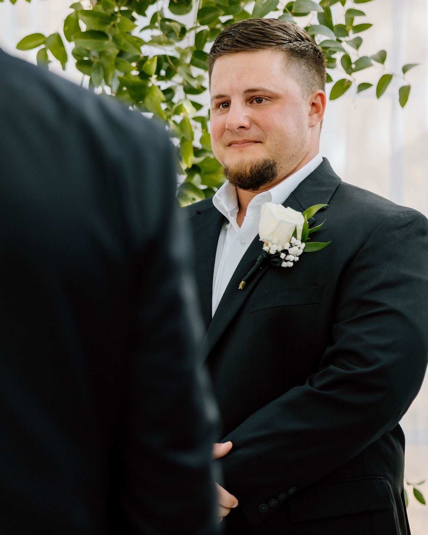 Groom in black suit with white rose boutonniere smiling at altar during wedding ceremony.
