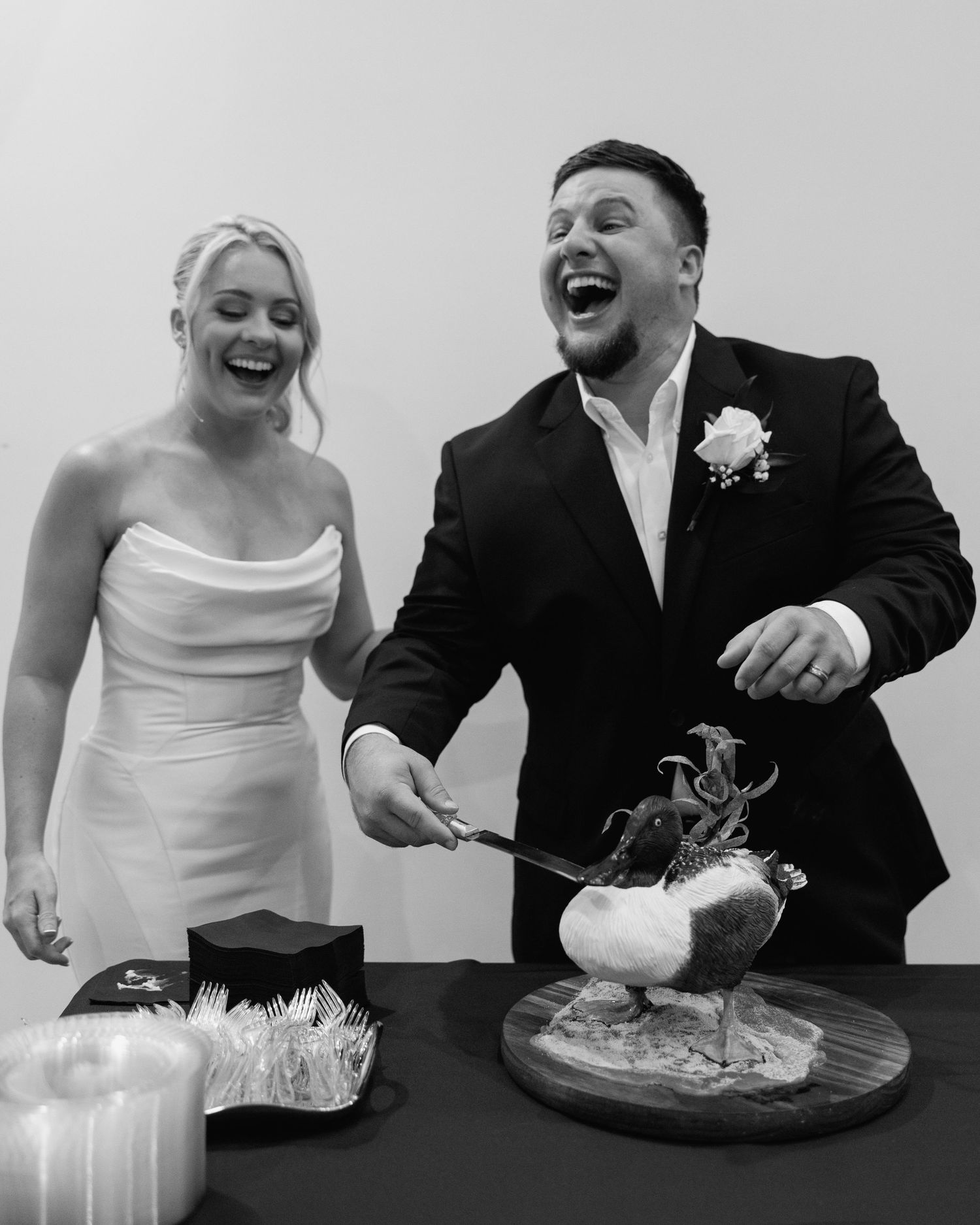 Black and white photo of laughing bride and groom cutting a decorative grooms cake at reception.