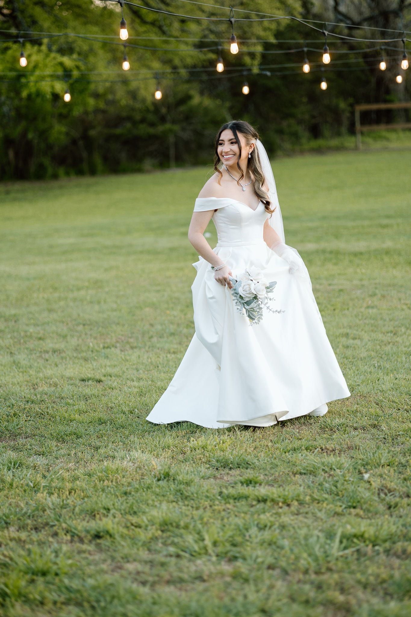 Bride walking on grass under string lights in classic off-shoulder wedding gown.