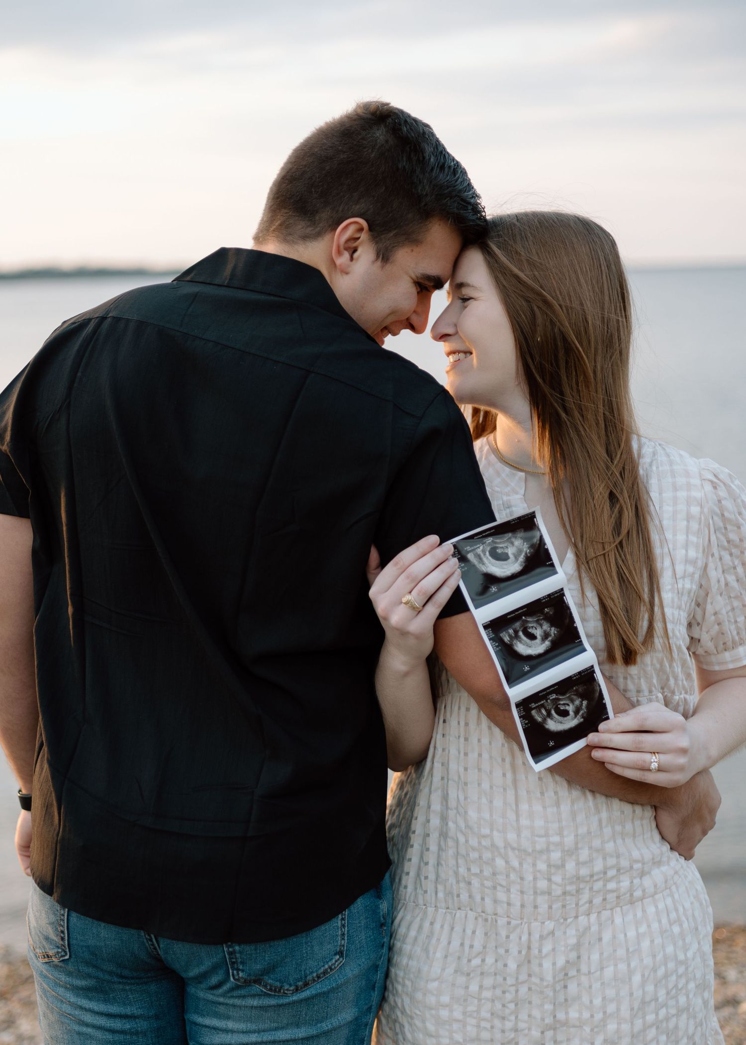 Couple touches foreheads in romantic moment while holding ultrasound photos by water.