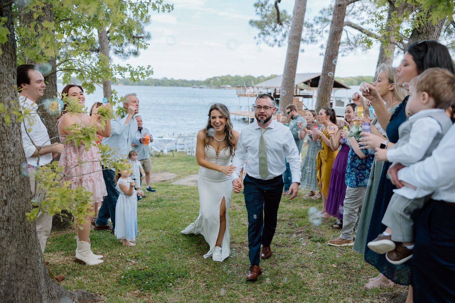 A couple walking down an aisle of guests on a grassy waterfront path.