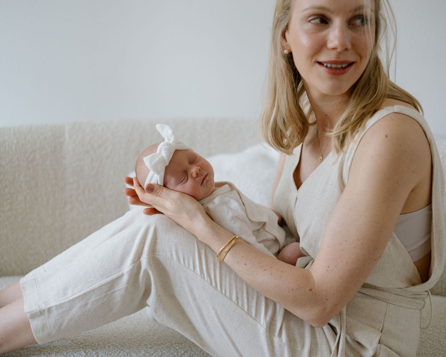 A parent cradles a sleeping newborn baby dressed in white on a light gray couch.