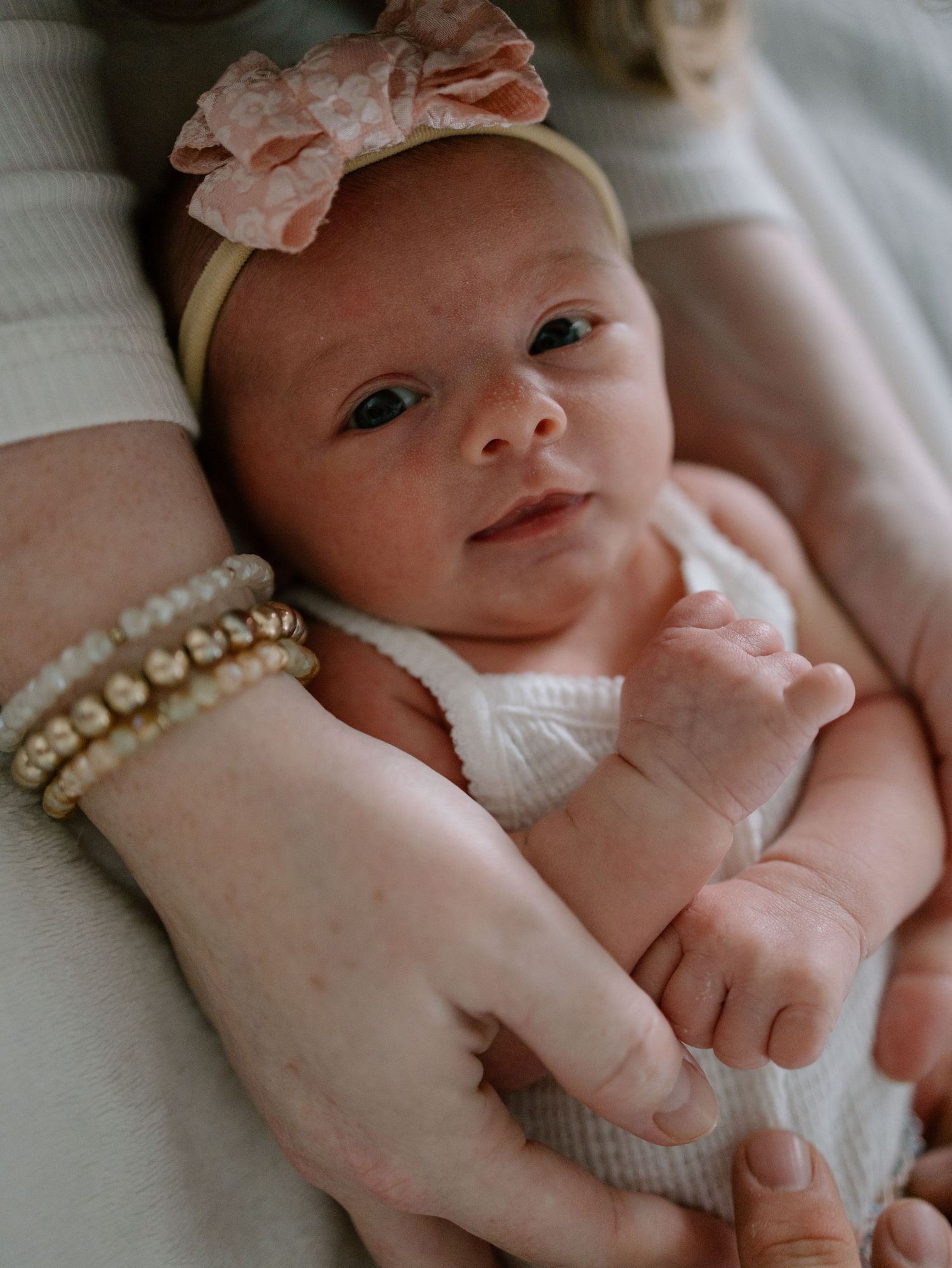 Baby wearing white outfit and peach floral headband lays on white blanket while adult hand with pearl bracelets touches arm.