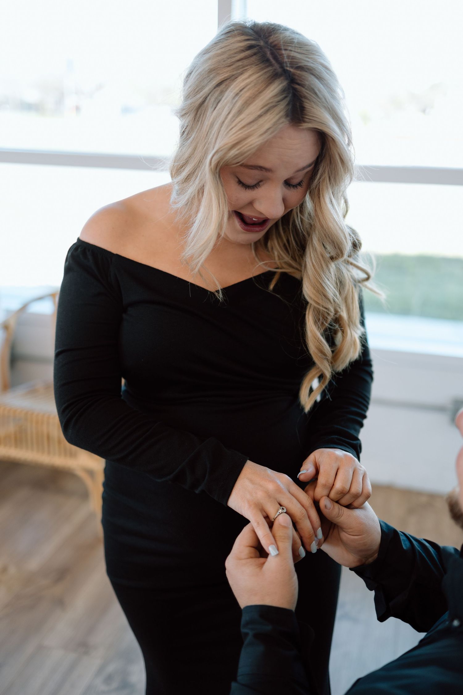 Woman in black off-shoulder dress smiles while someone places ring on her finger during proposal moment.