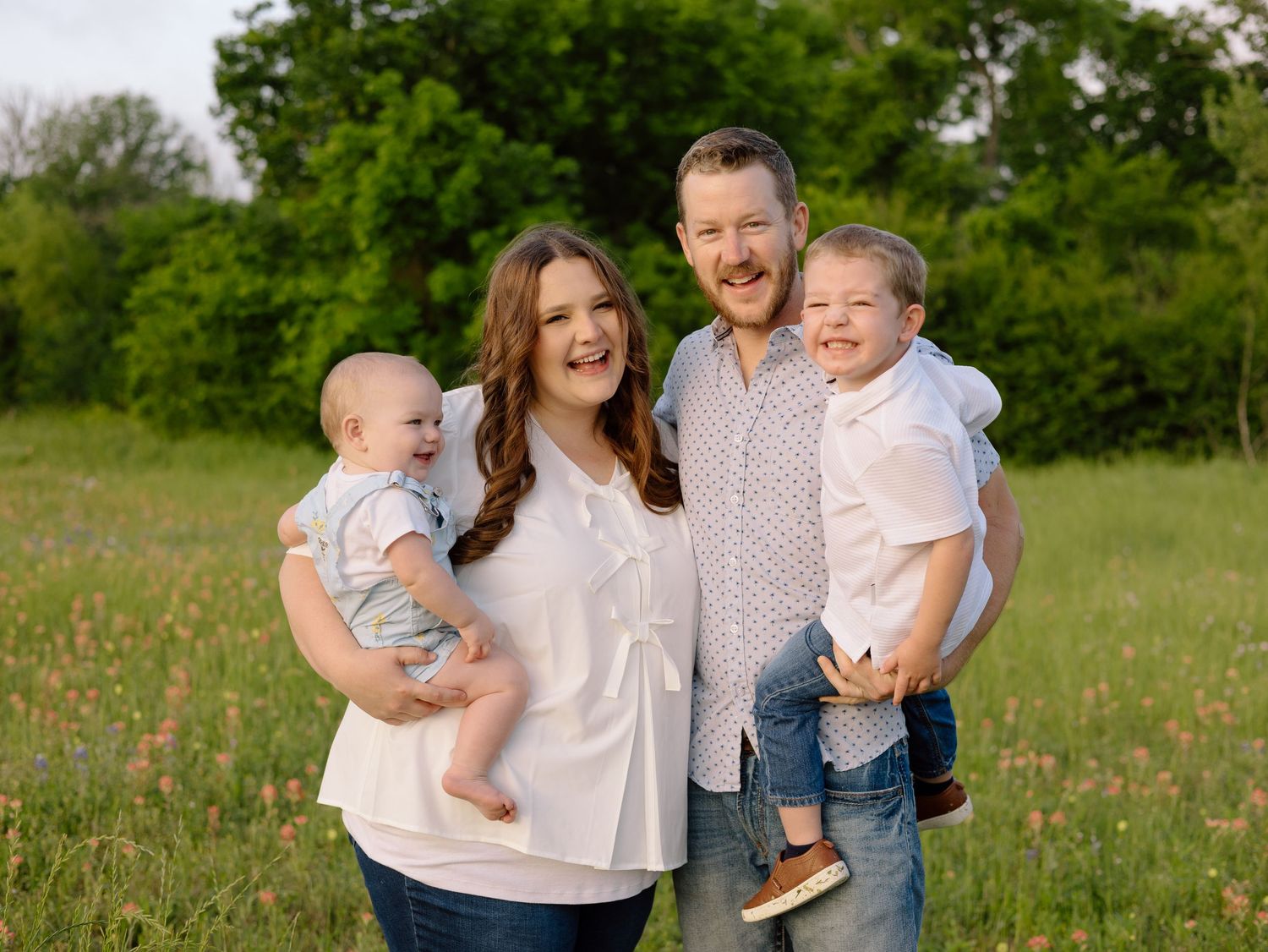 Family of four poses together outdoors in a field with green trees and grass in the background.