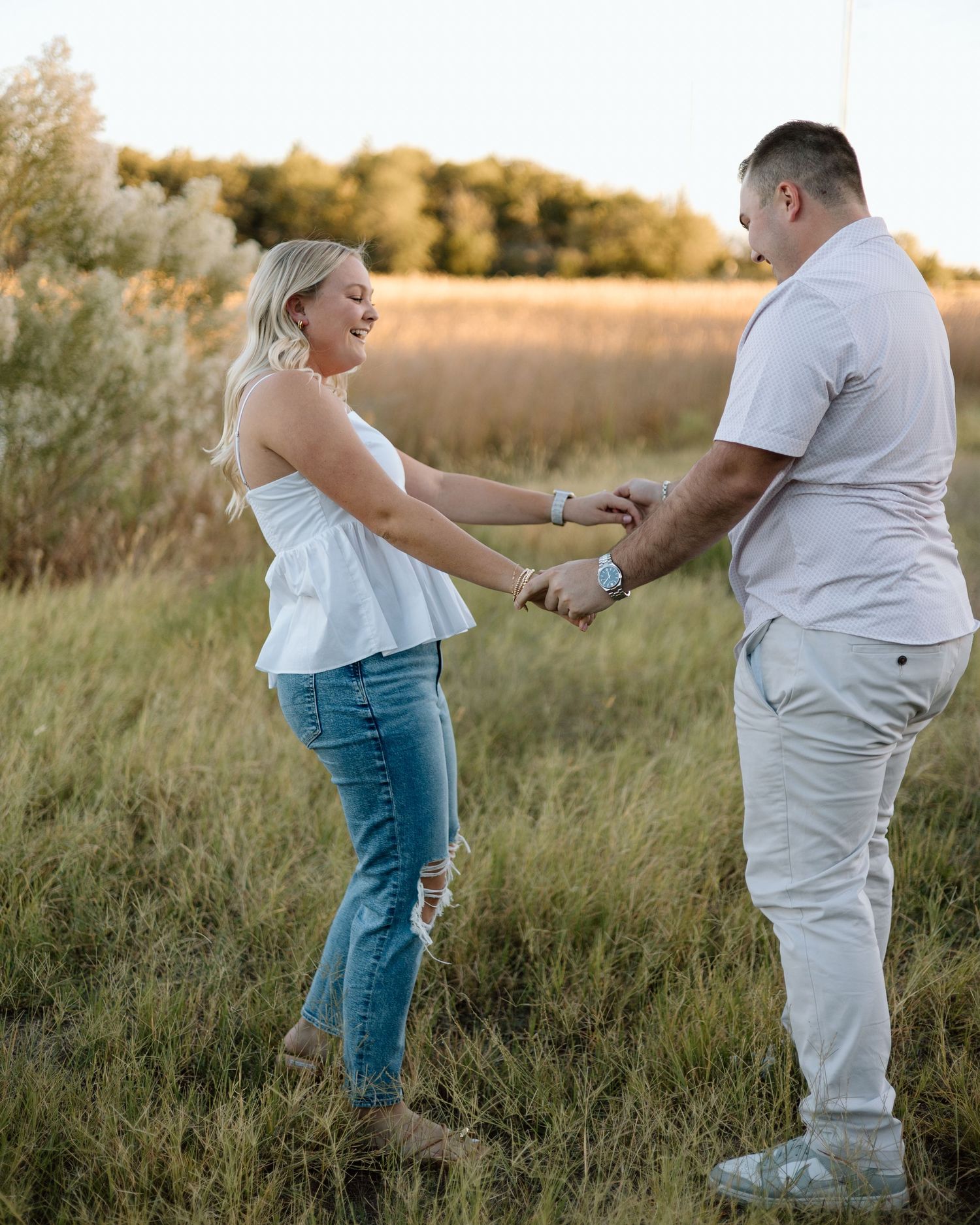Couple holding hands and dancing in a golden field at sunset, woman in white top and jeans, man in white outfit.