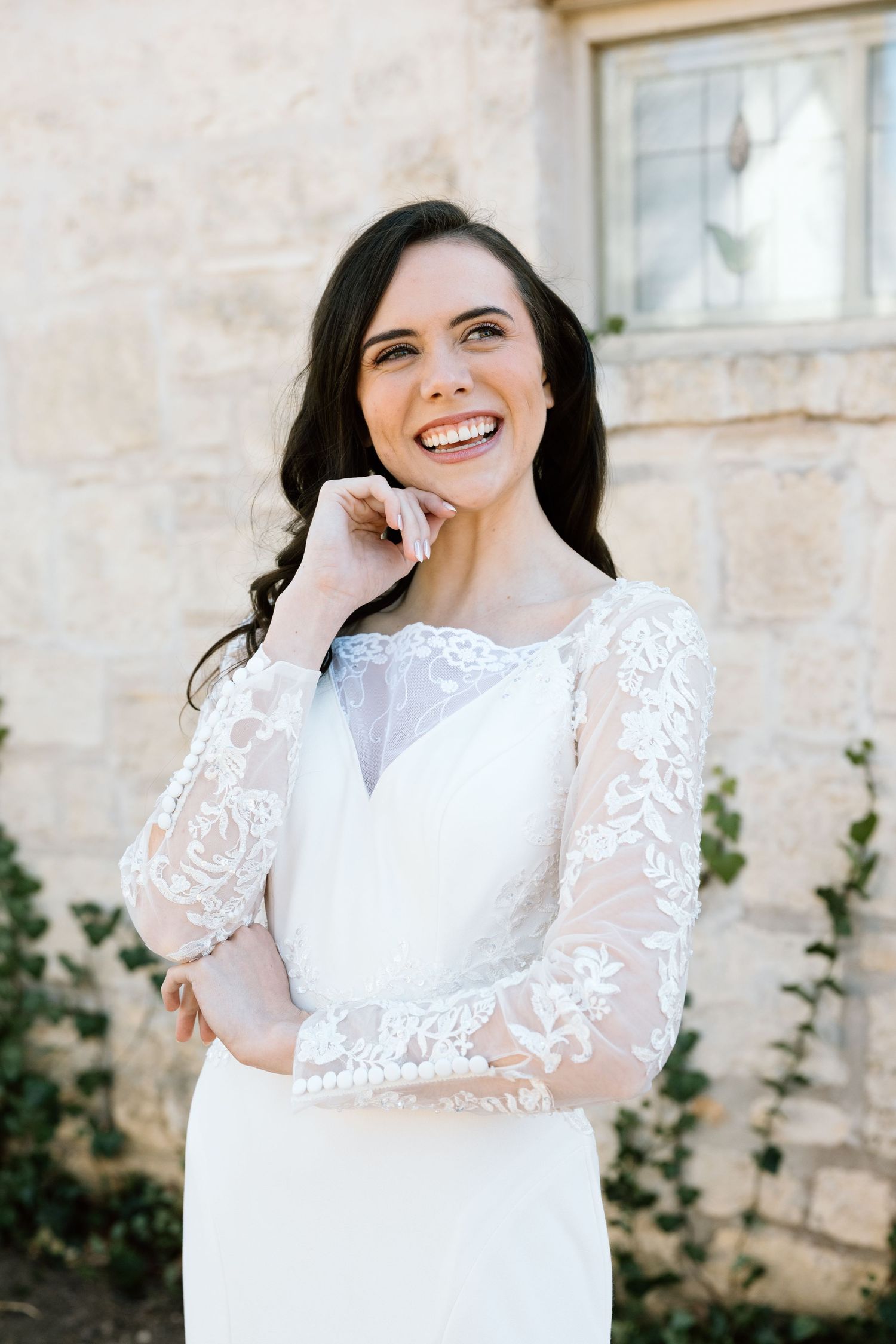 Smiling bride in long-sleeve lace wedding dress standing before stone wall.