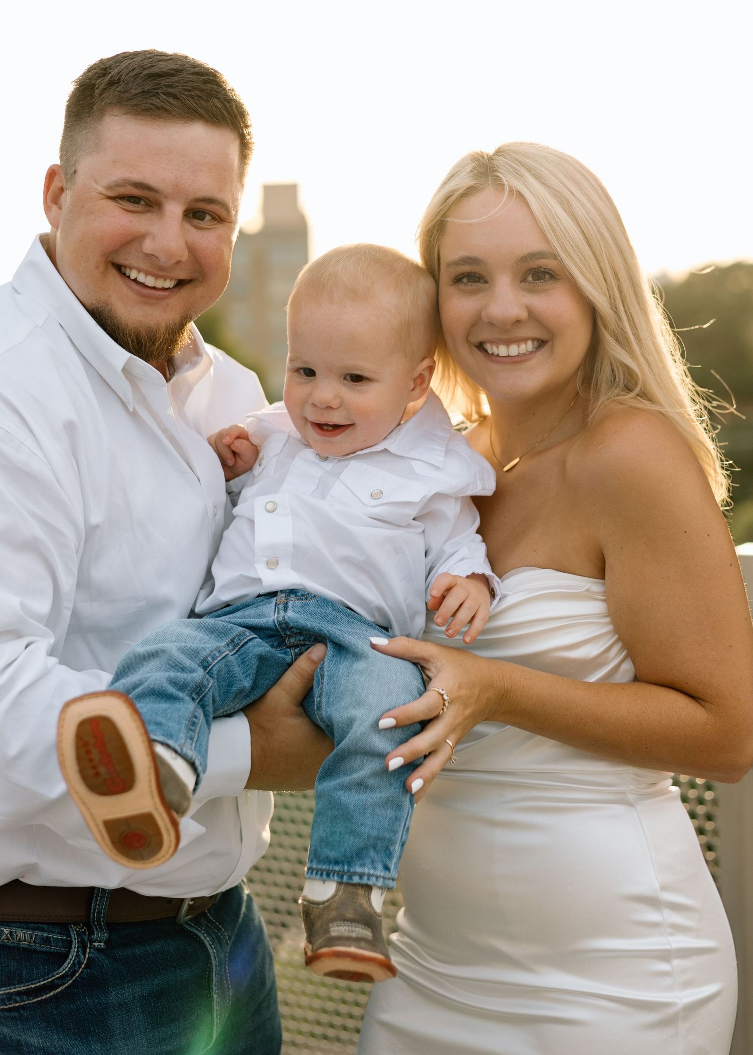 Happy family portrait with parents in white outfits holding smiling baby boy outdoors in golden hour light.