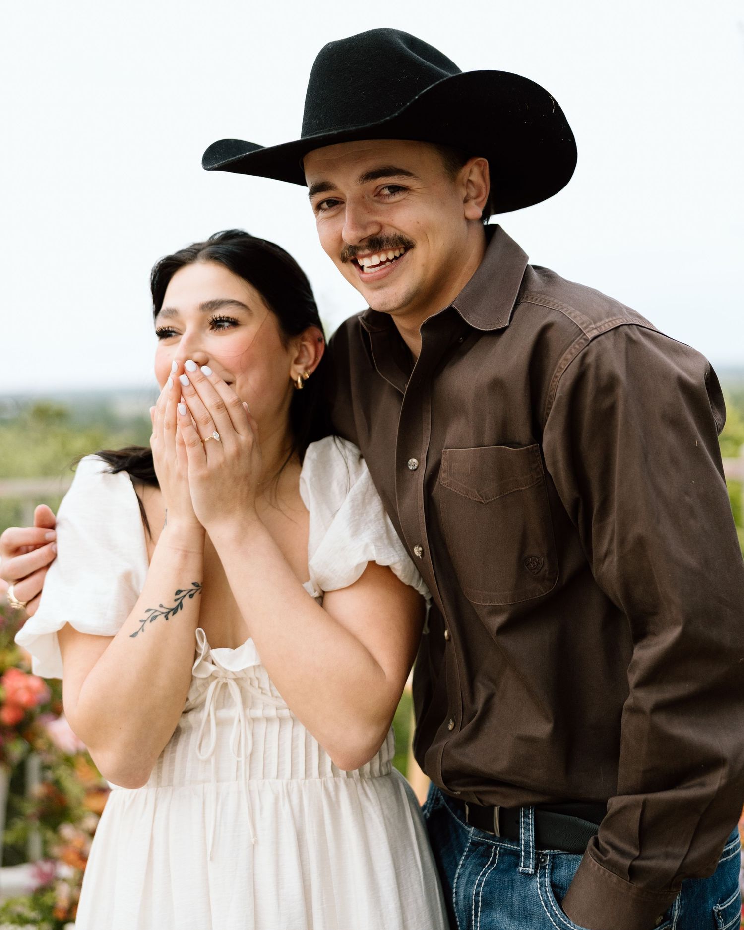 A smiling couple poses outdoors, woman in white dress covering her mouth, man in brown shirt and black cowboy hat.