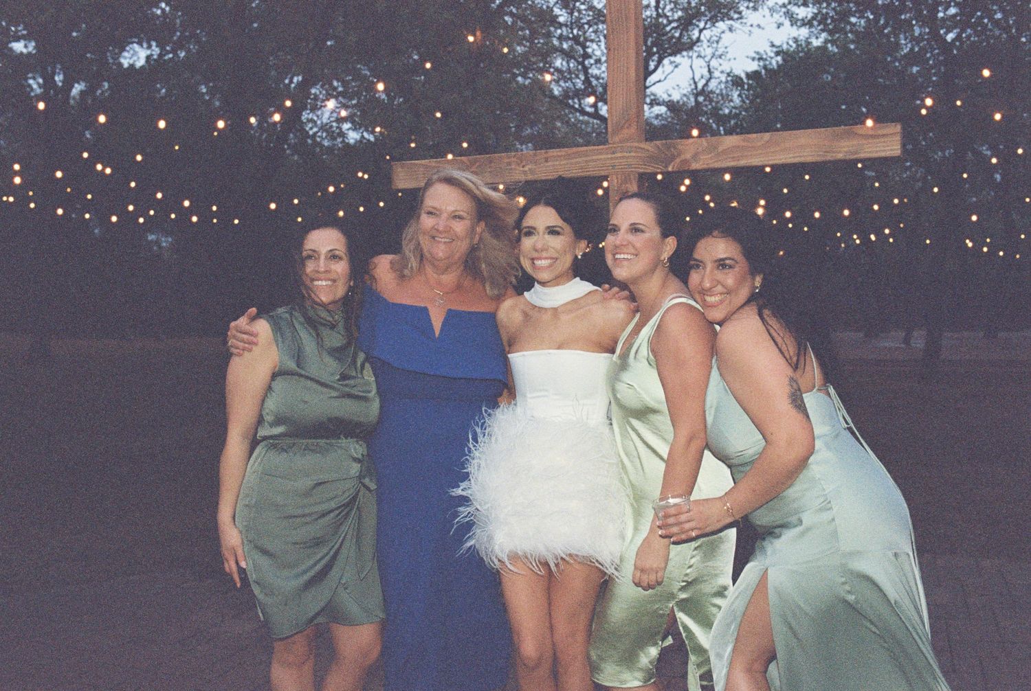 Wedding party members in coordinated dresses pose together under string lights at an outdoor venue.