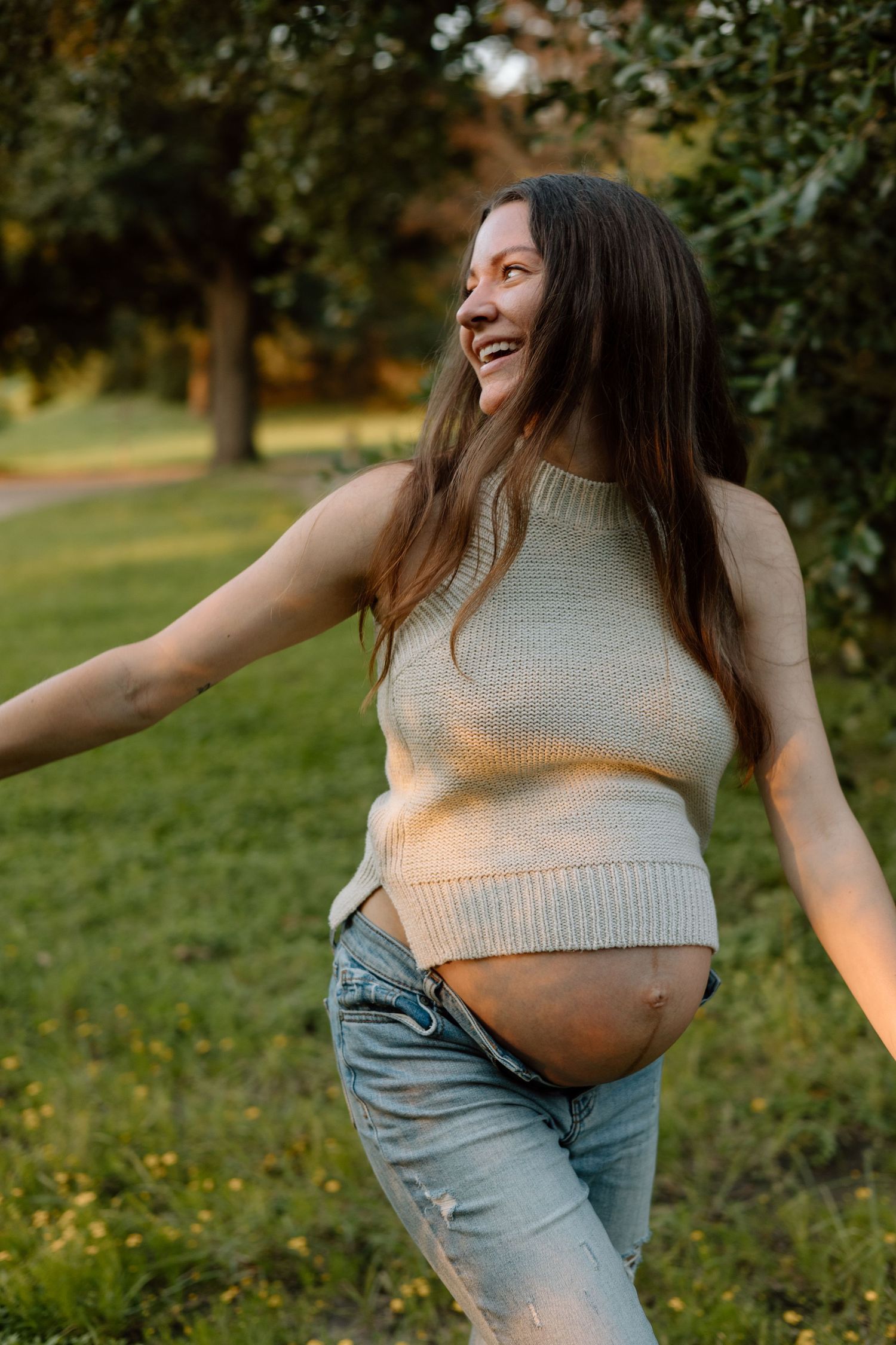 A pregnant woman in a cream sweater poses playfully in a park at sunset.