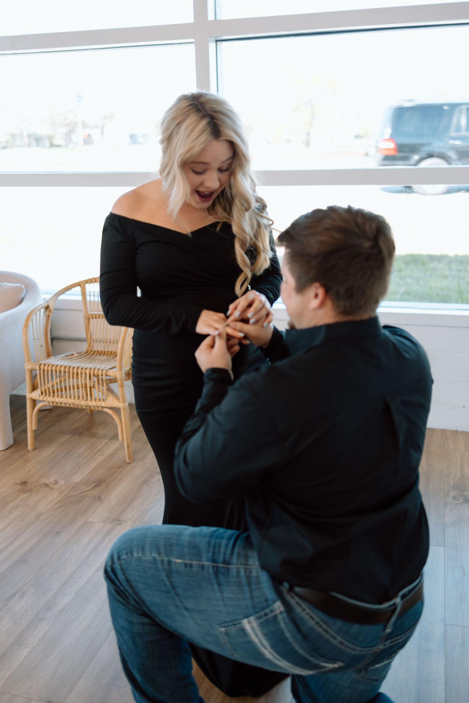 A marriage proposal moment captured indoors with someone in a black off-shoulder dress and another kneeling on hardwood flooring.