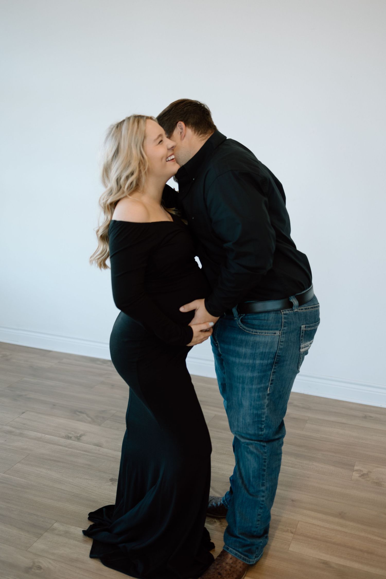A tender maternity photoshoot features a couple in black attire sharing an embrace on wooden flooring.