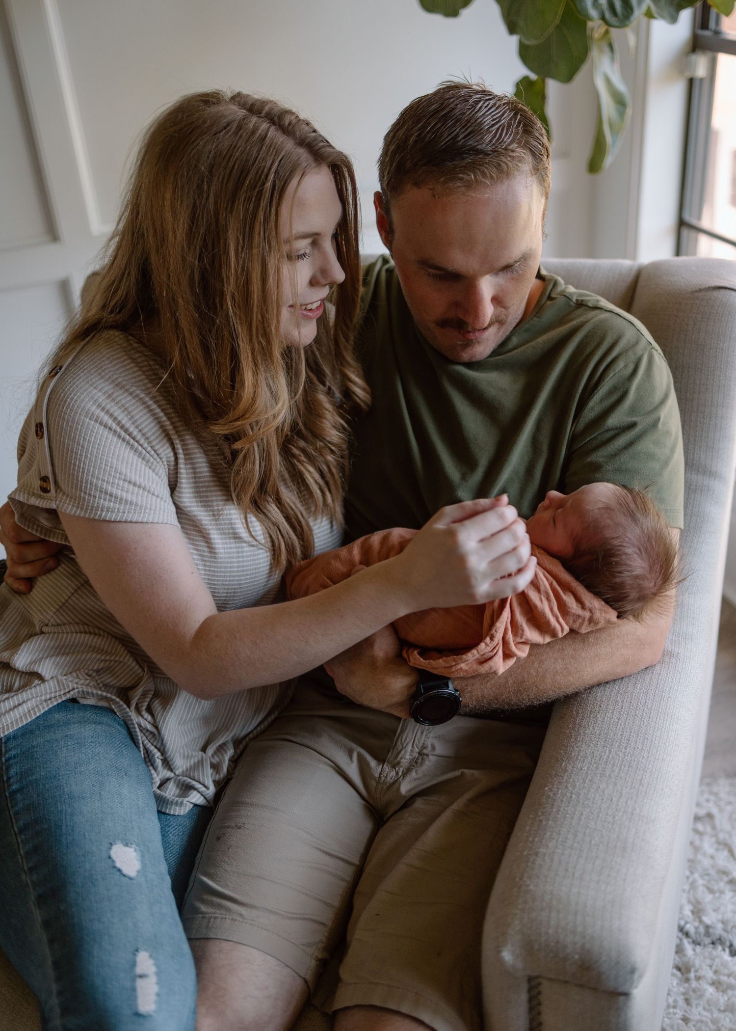 A loving couple sits together on a grey couch holding a newborn baby in an orange blanket by a sunny window.