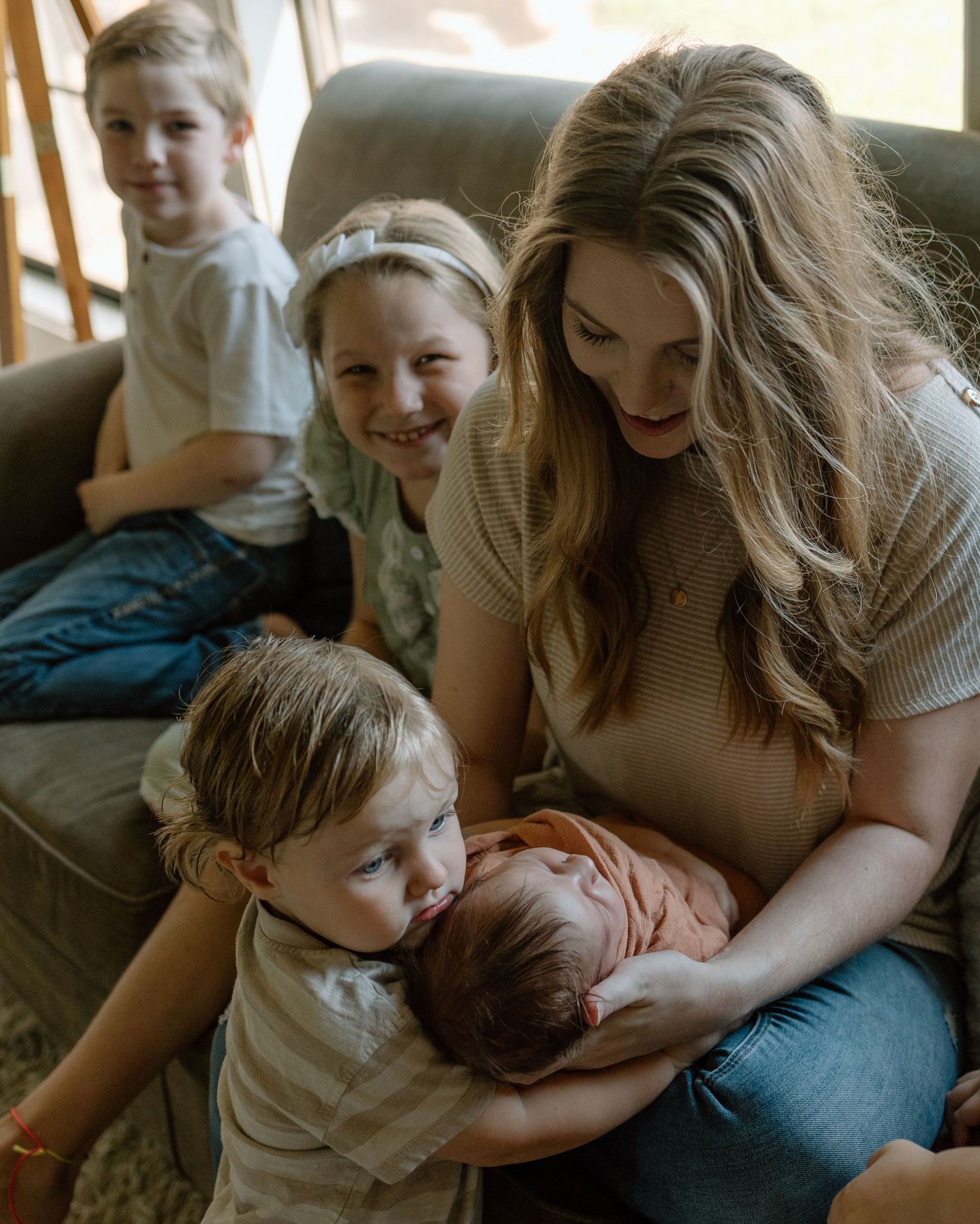Family sits together on sofa as parent holds newborn baby while older siblings gather close in candid moment.
