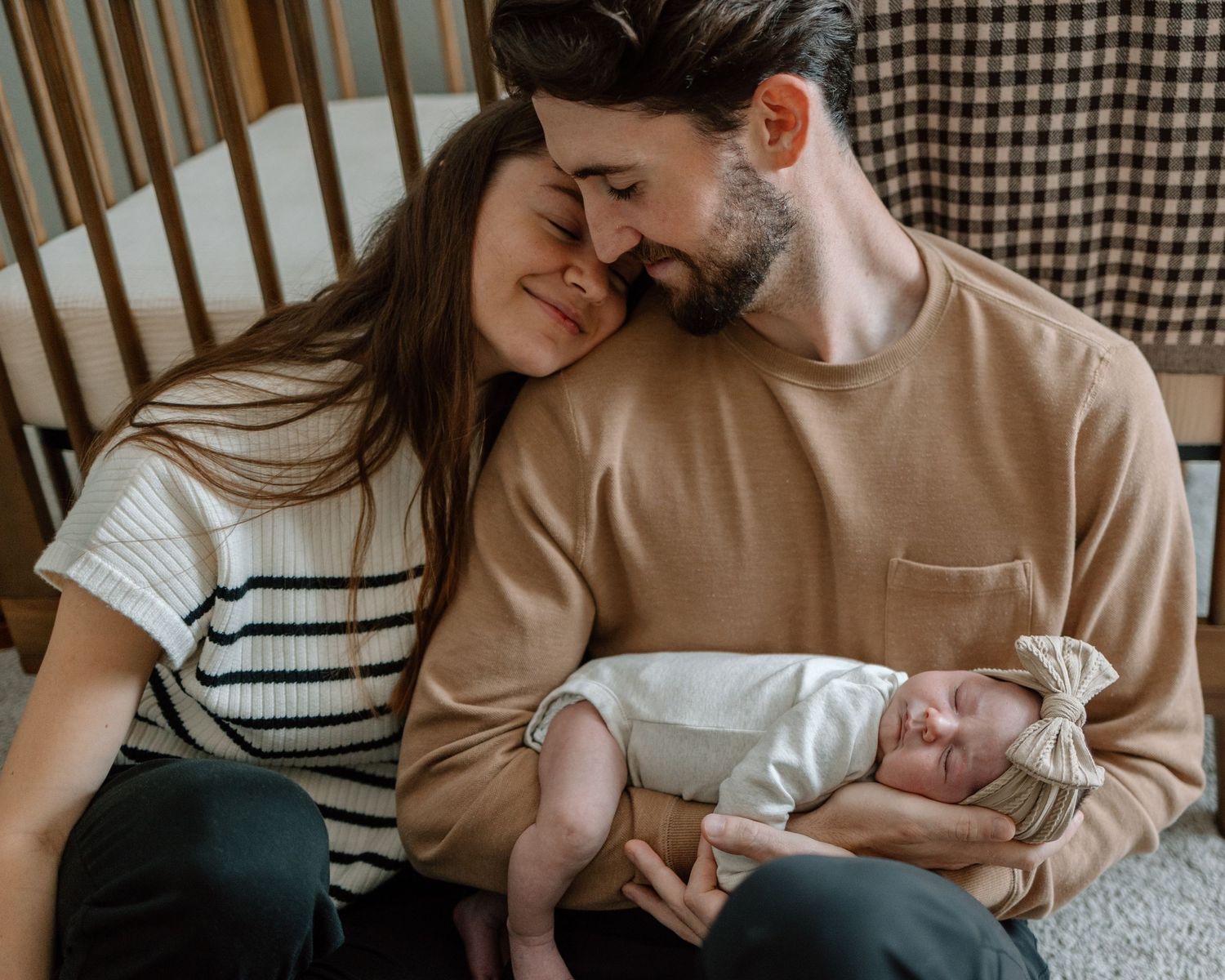 Family of three sharing tender moment while sitting by wooden crib with plaid backdrop.
