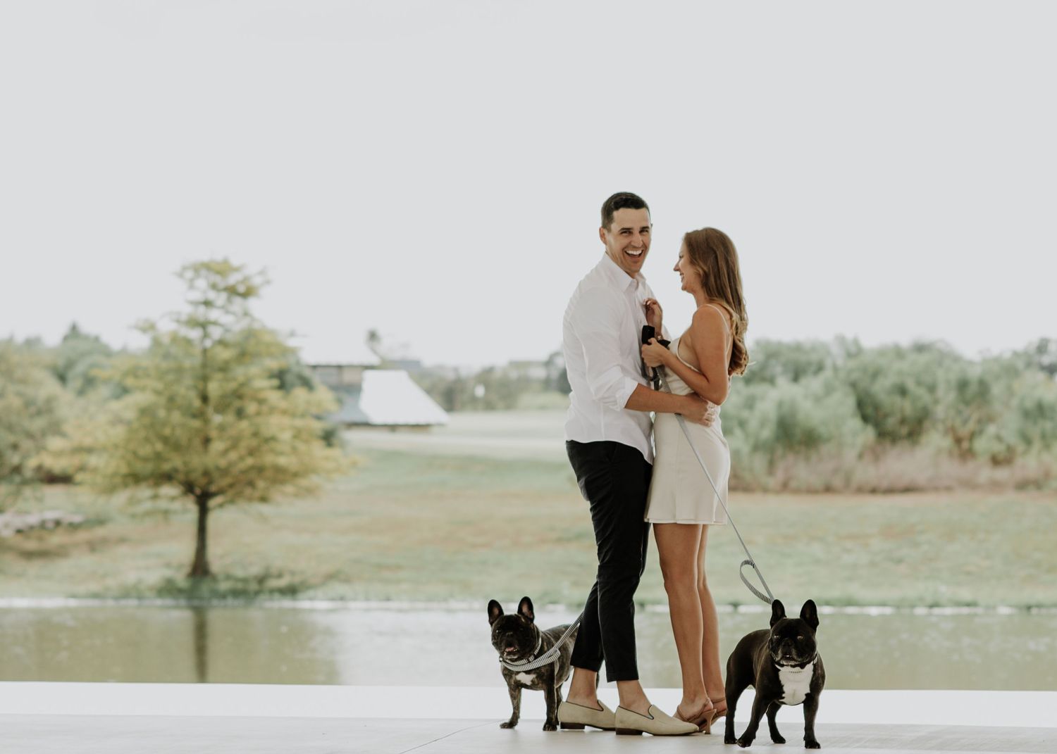 A couple stands together on a concrete path with two black dogs beside them.
