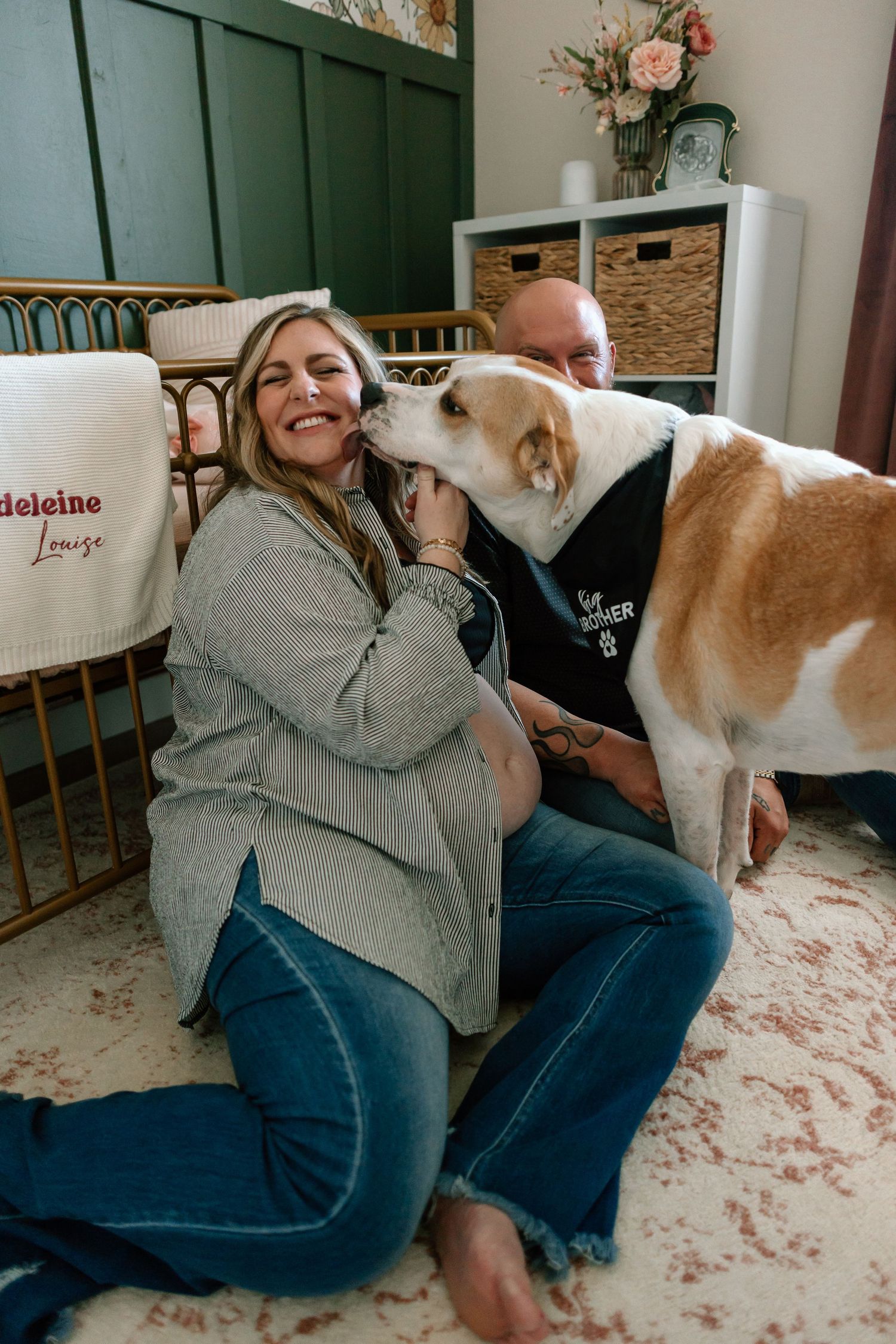 Dog gives kisses to expecting mother while couple relaxes on floor at home.