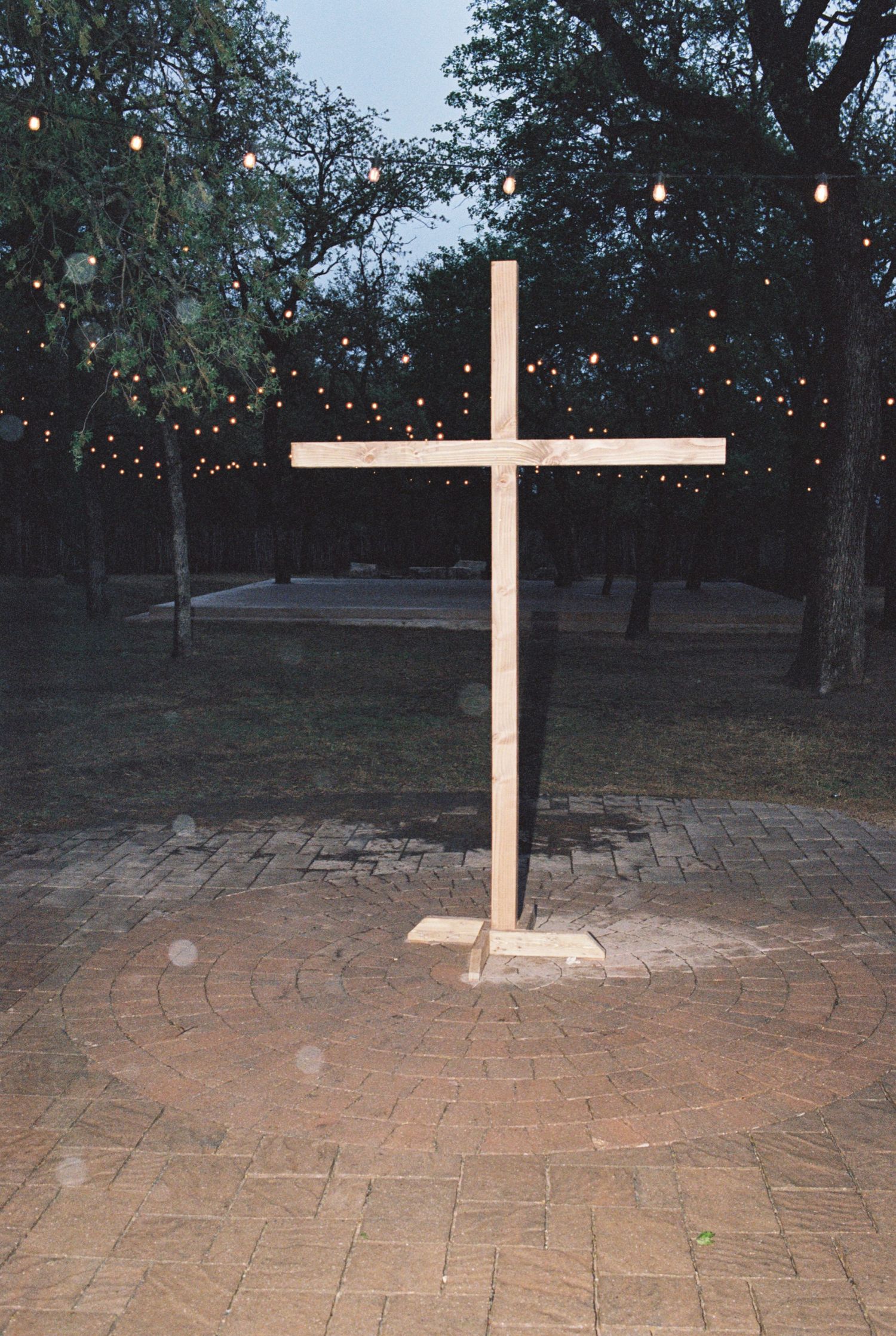 A white illuminated cross stands on brick pavement with string lights and trees in the background.
