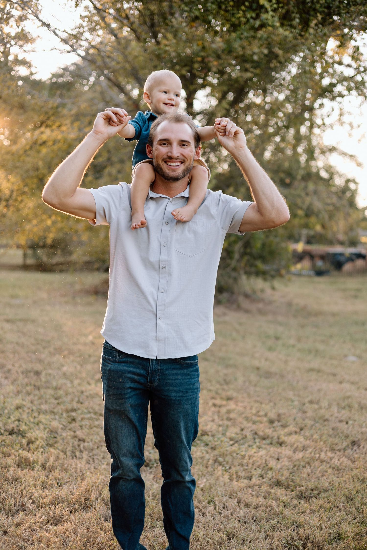 A smiling father carries his baby on his shoulders in a sunny outdoor park setting.