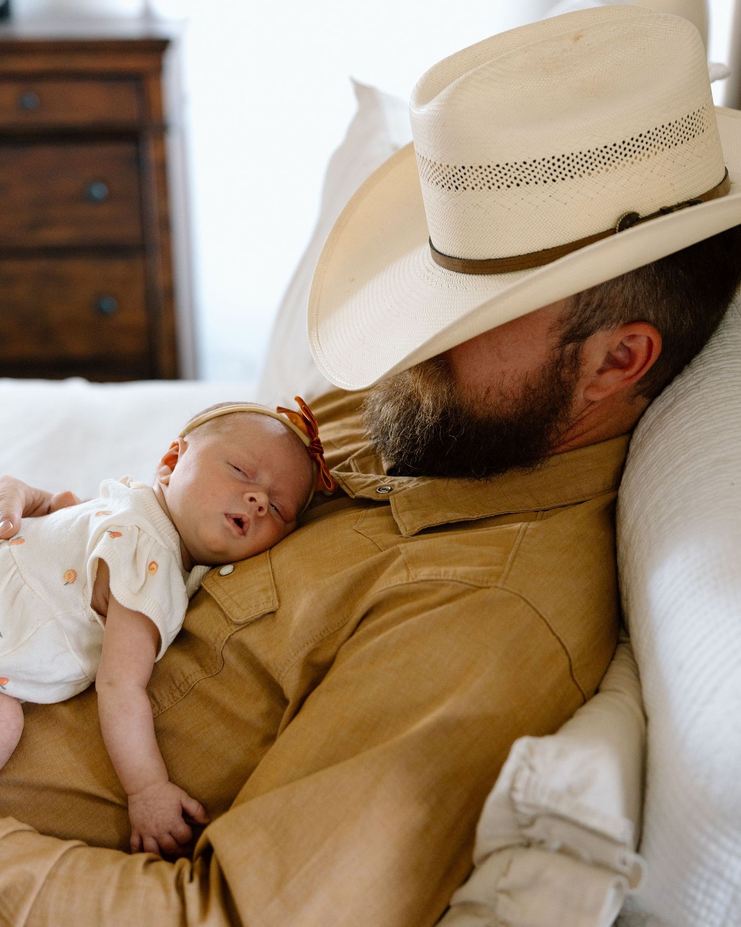 Bearded father in cowboy hat and tan shirt rests with sleeping newborn baby on his chest.