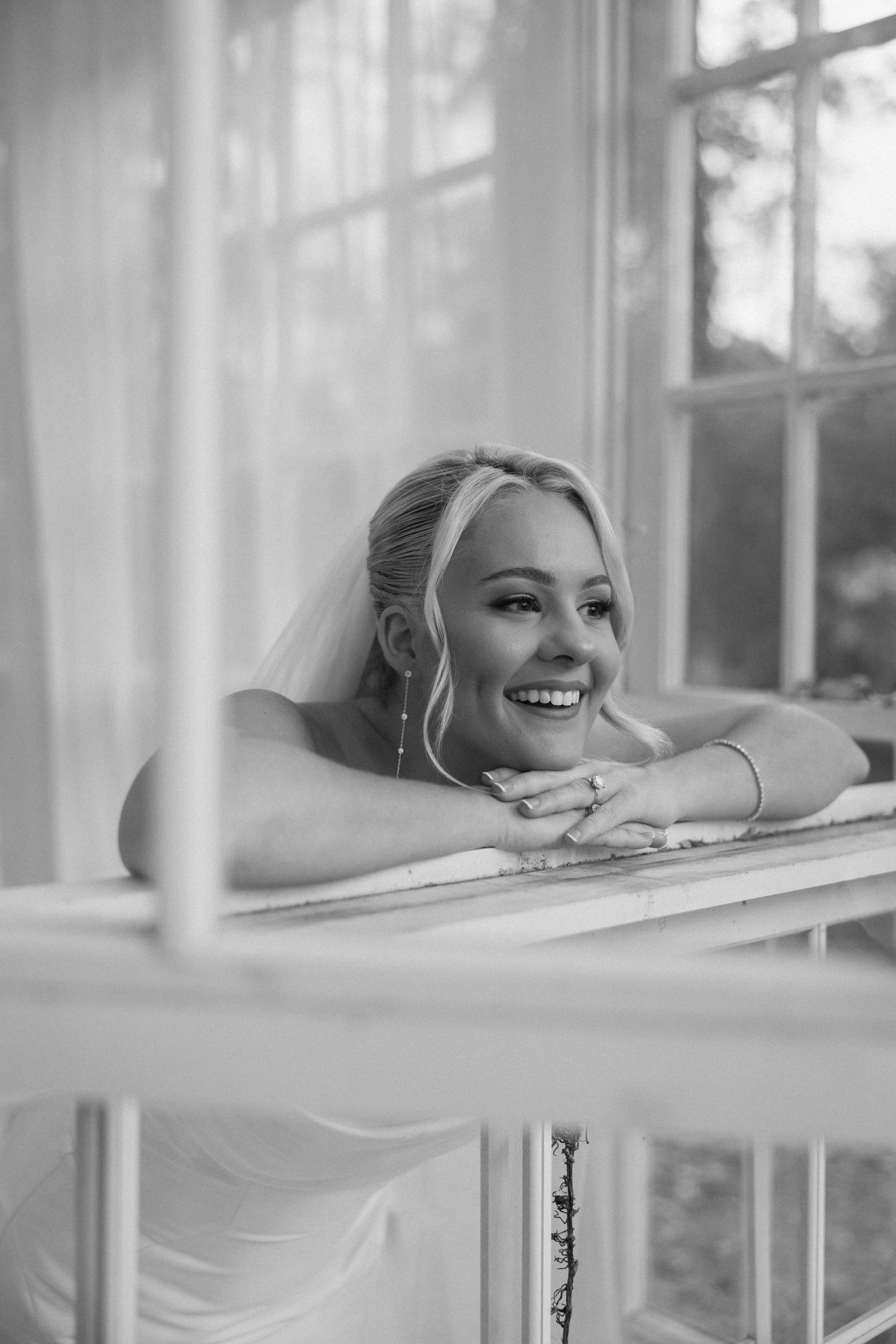 Smiling bride in veil leans on windowsill in black and white portrait.